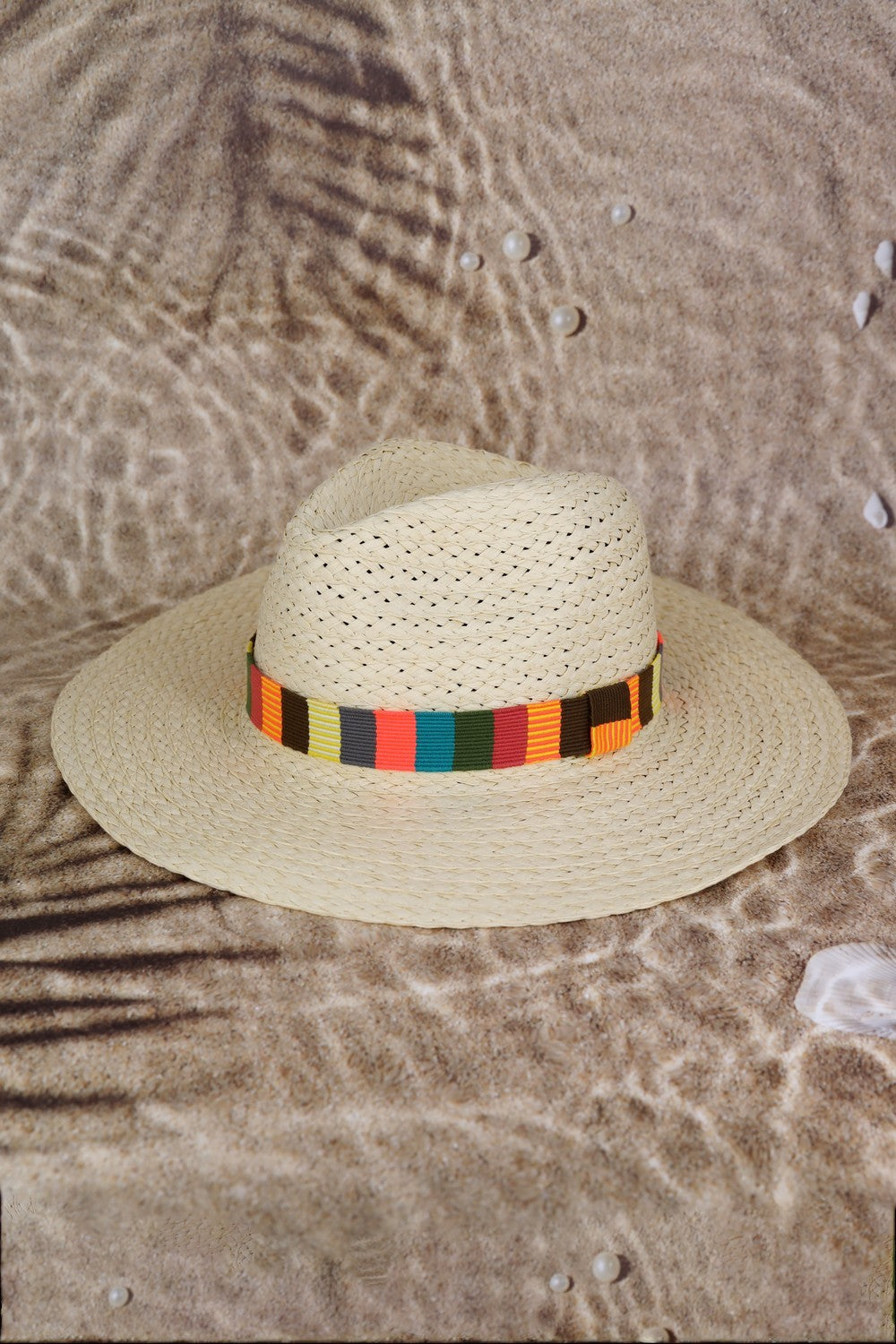 The Fame Contrast Wide Brim Straw Hat, featuring a vibrant multicolor band, is elegantly showcased on a sandy beach background with delicate palm leaf shadows, making it the ideal stylish accessory for sun protection.