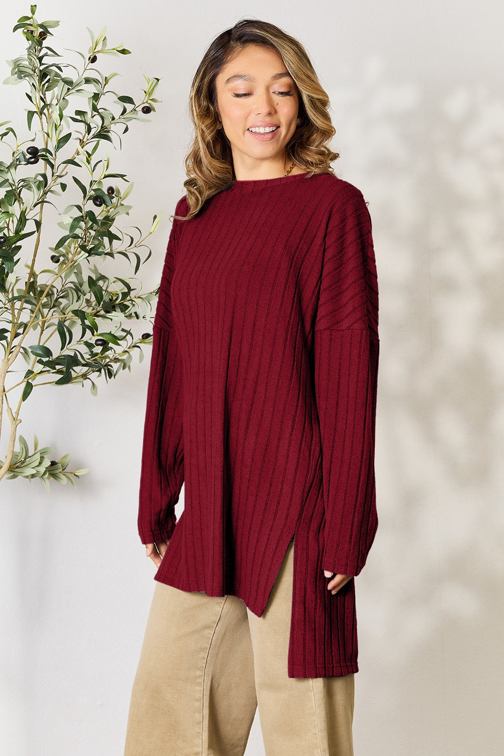 A person with shoulder-length hair smiles while wearing a beige ribbed sweater called the Basic Bae Full Size Ribbed Round Neck Long Sleeve Slit Top, along with matching beige pants, showcasing a basic style. They stand next to a green plant.