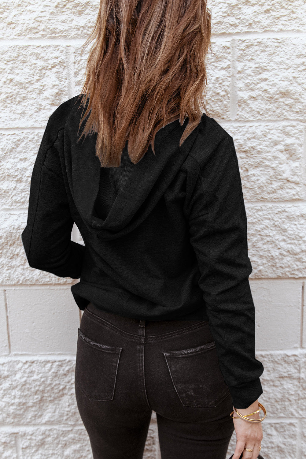 A person with long hair facing away from the camera, wearing a Black Casual Solid Color Lace-up Hoodie and black jeans, standing in front of a white brick wall, epitomizing fashionable comfort.