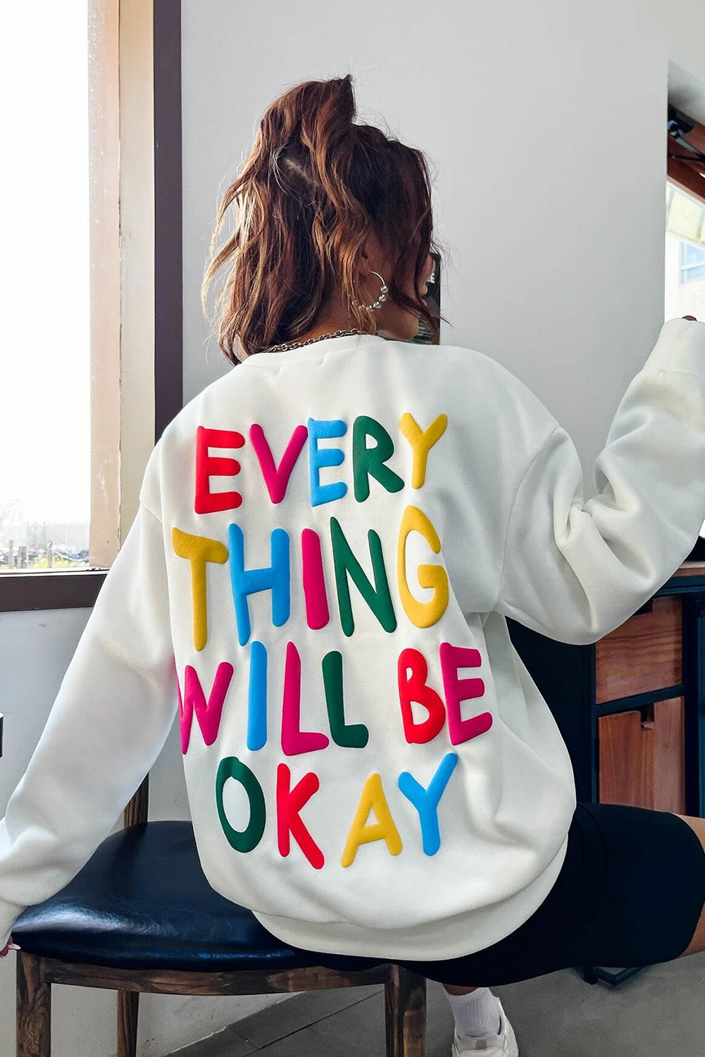 A person wearing a white beanie, the EVERY THING WILL BE OKAY Colorful Letters Sweatshirt, and sunglasses, holding a coffee cup, stands outside near windows.