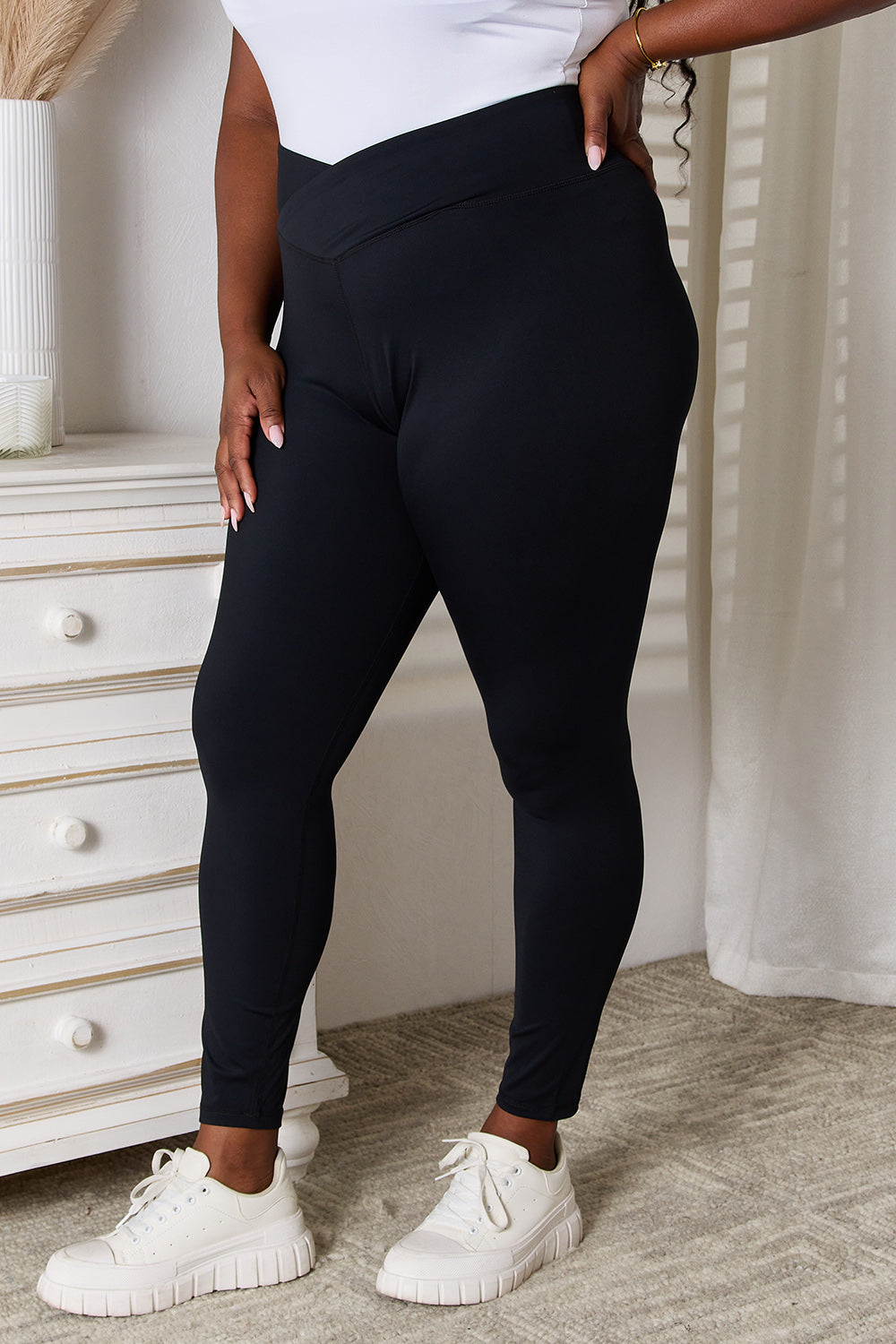 A person is standing in a room wearing Basic Bae V-Waistband Sports Leggings that offer a flattering fit, paired with white slip-on shoes. To the left, there is a white dresser with drawers. This outfit highlights an active lifestyle, seamlessly blending comfort and style.