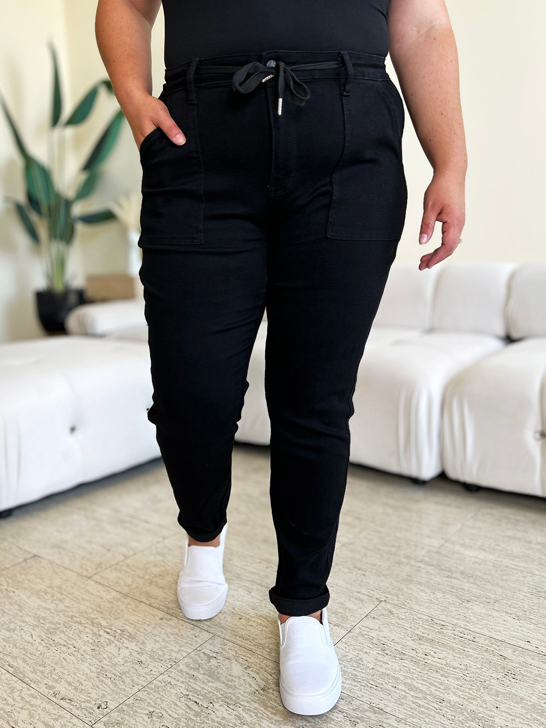 Person standing indoors, wearing a white top, black Judy Blue Full Size High Waist Double Roll Cuff Jeans, and white slip-on shoes, next to white couches and a plant in the background.