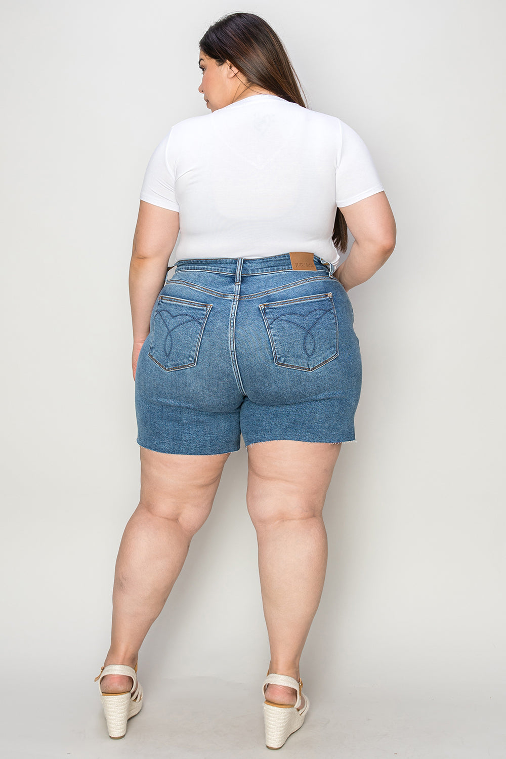 A woman in a white short-sleeve top and Judy Blue Full Size High Waist Slim Denim Shorts stands against a plain backdrop. She is smiling with her hands resting on her hips, wearing white open-toe wedge sandals that emphasize the quality denim fabric of her high-waisted design.