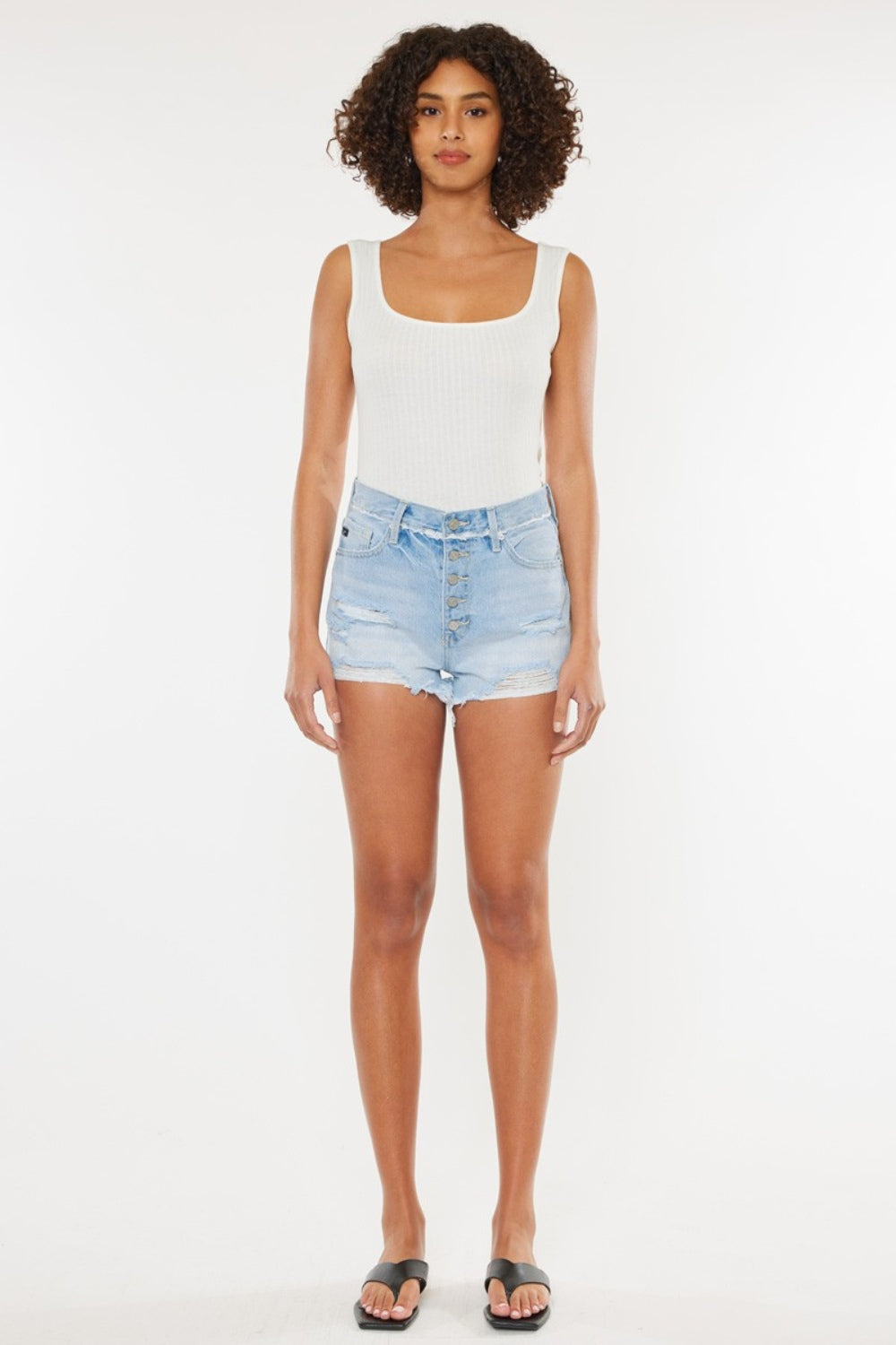 A person wearing a white tank top and Kancan Distressed Button Fly Denim Shorts poses against a plain background, showcasing a quintessential summer wardrobe staple.