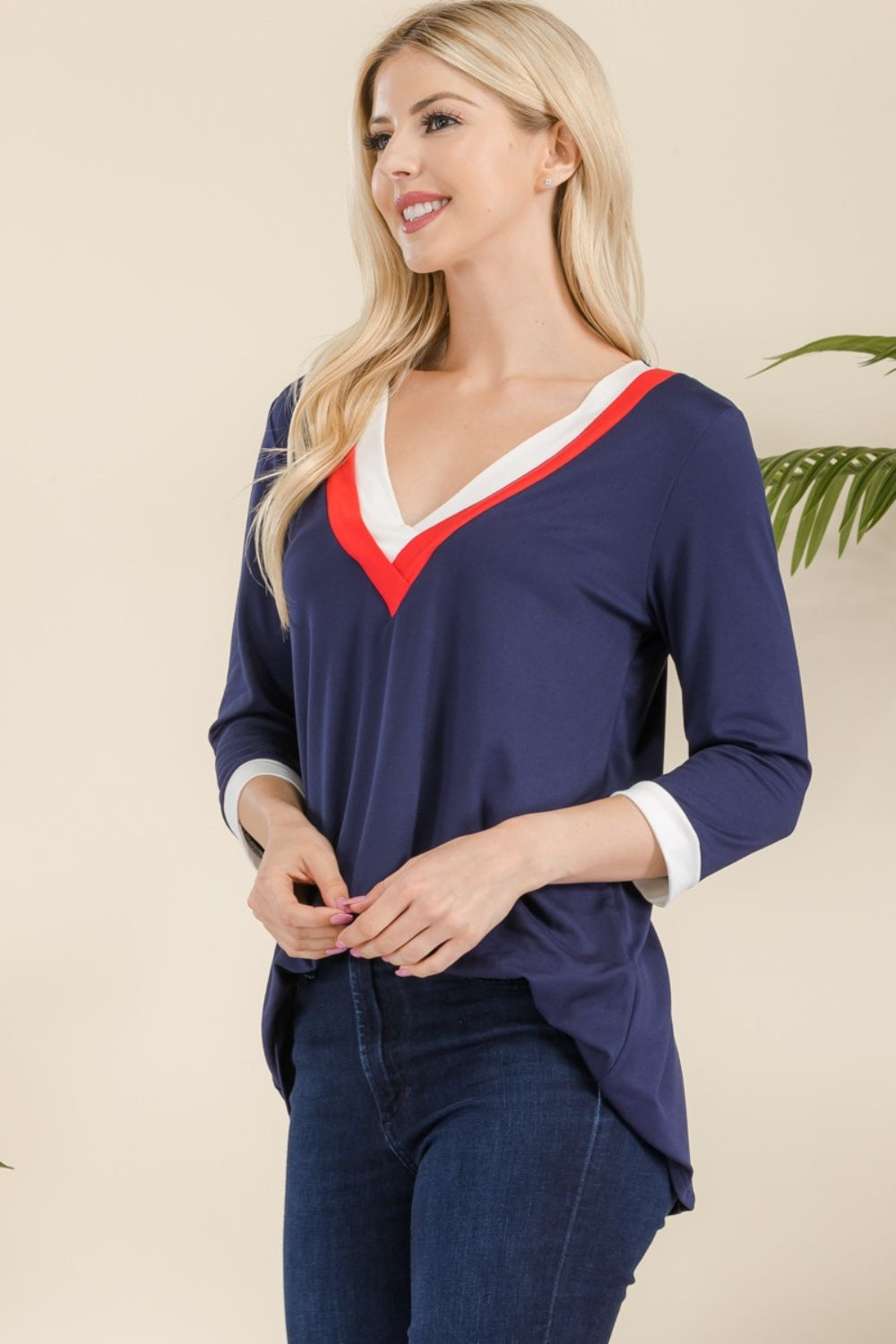 A woman with long blonde hair wearing the Celeste Full Size V-Neck Contrast T-Shirt crafted from lightweight material and dark blue jeans stands smiling.