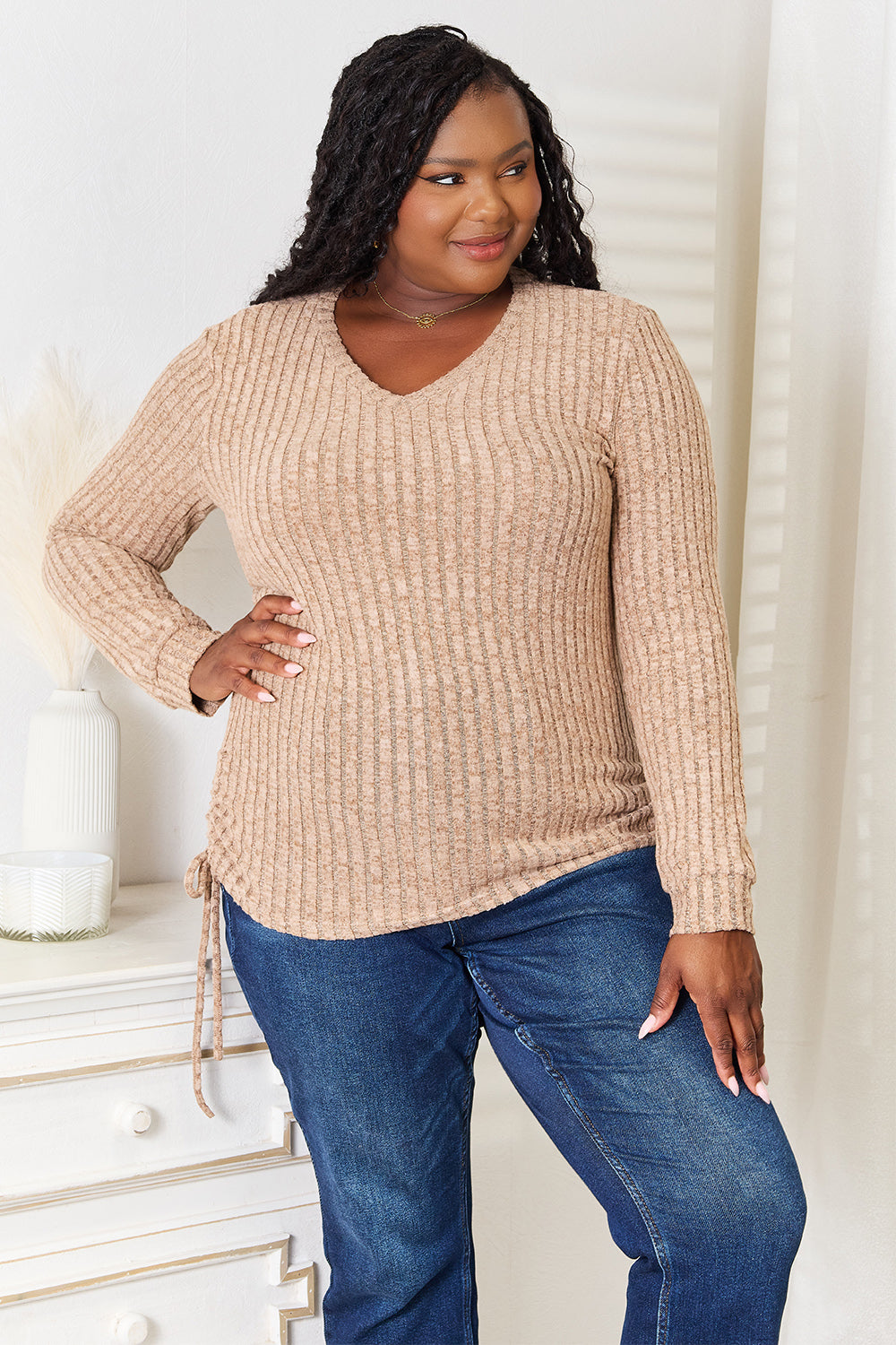 A woman stands indoors against a neutral background, wearing the "Double Take Drawstring Ribbed Long Sleeve T-Shirt" in light purple paired with dark pants. She is smiling and posing with one hand on her hip.