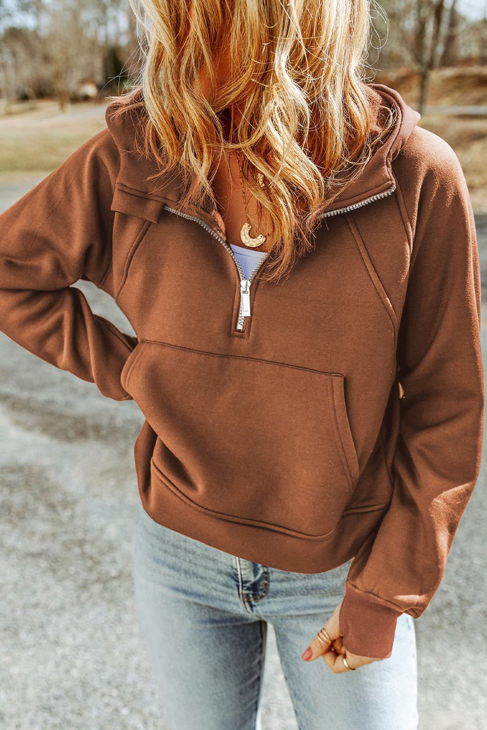 A person with long hair is walking away wearing a Brown Quarter Zip Kangaroo Pocket Hoodie and light blue ripped jeans. The background shows an outdoor setting.