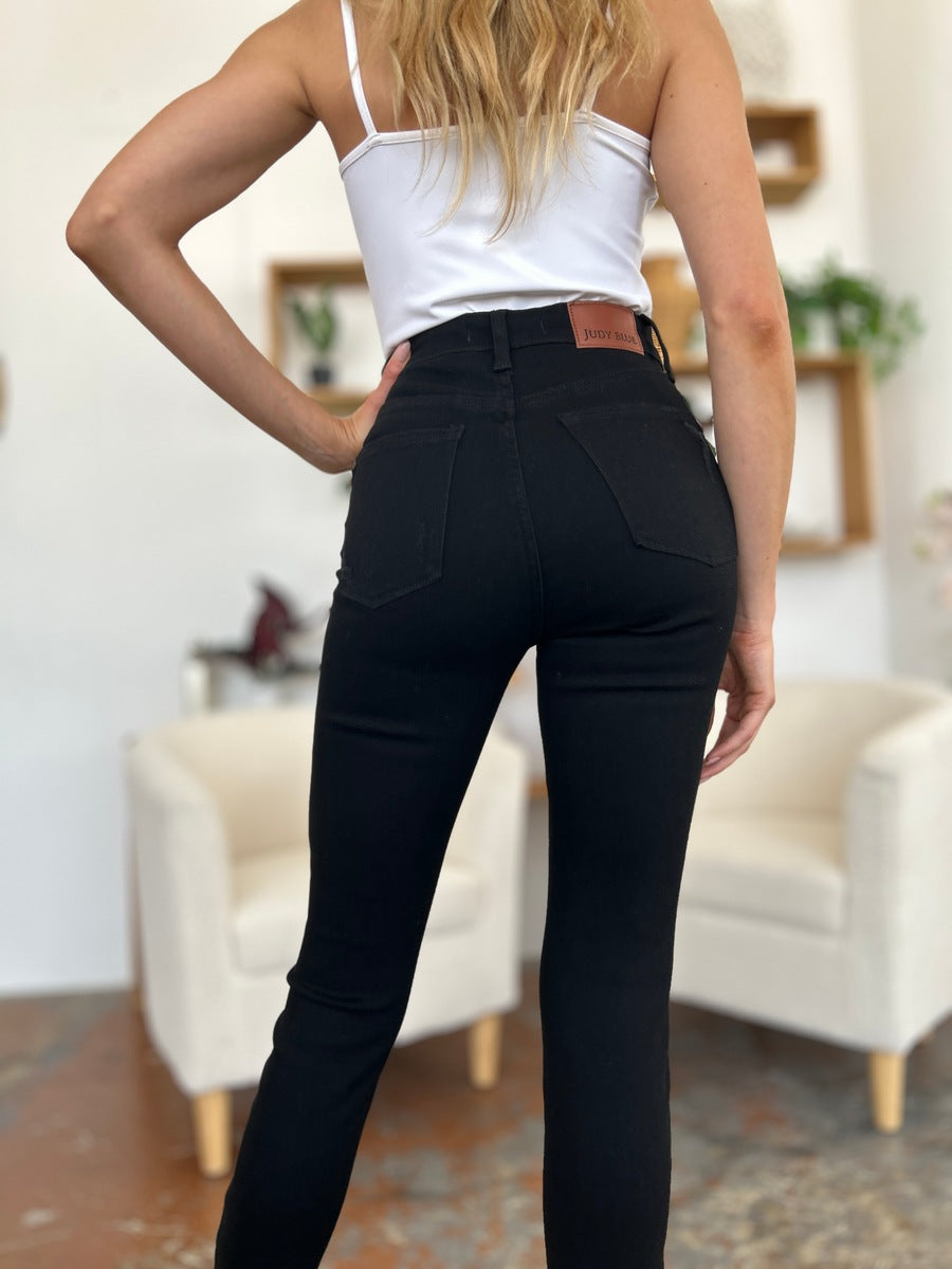 A person wearing a white T-shirt and Judy Blue Full Size Distressed Tummy Control High Waist Skinny Jeans poses barefoot in a room with white chairs and decorative plants in the background.