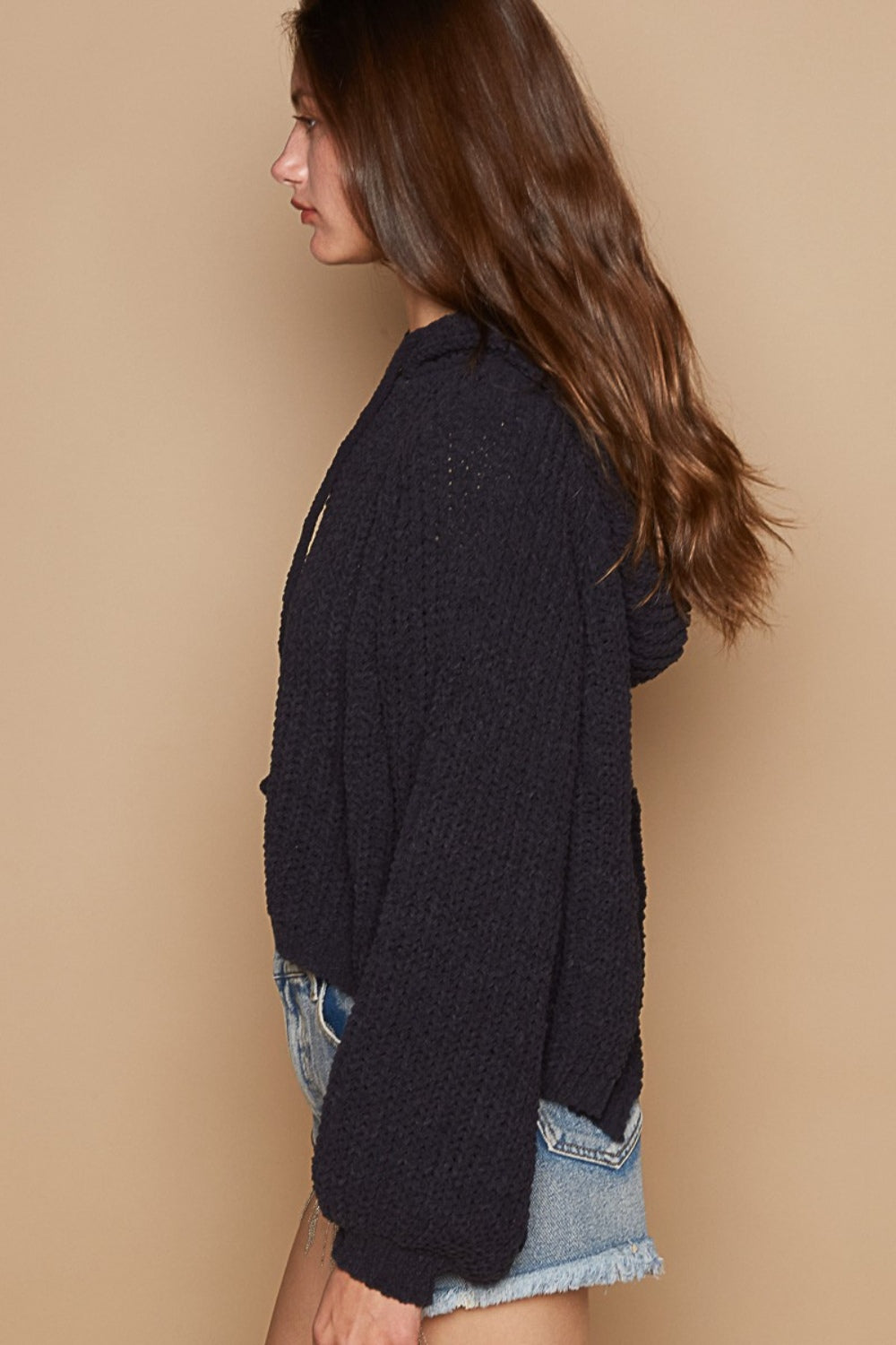 A person wearing a POL Back Open Slit Balloon Sleeve Crop Hooded Sweater in black and distressed blue jeans stands against a beige background.