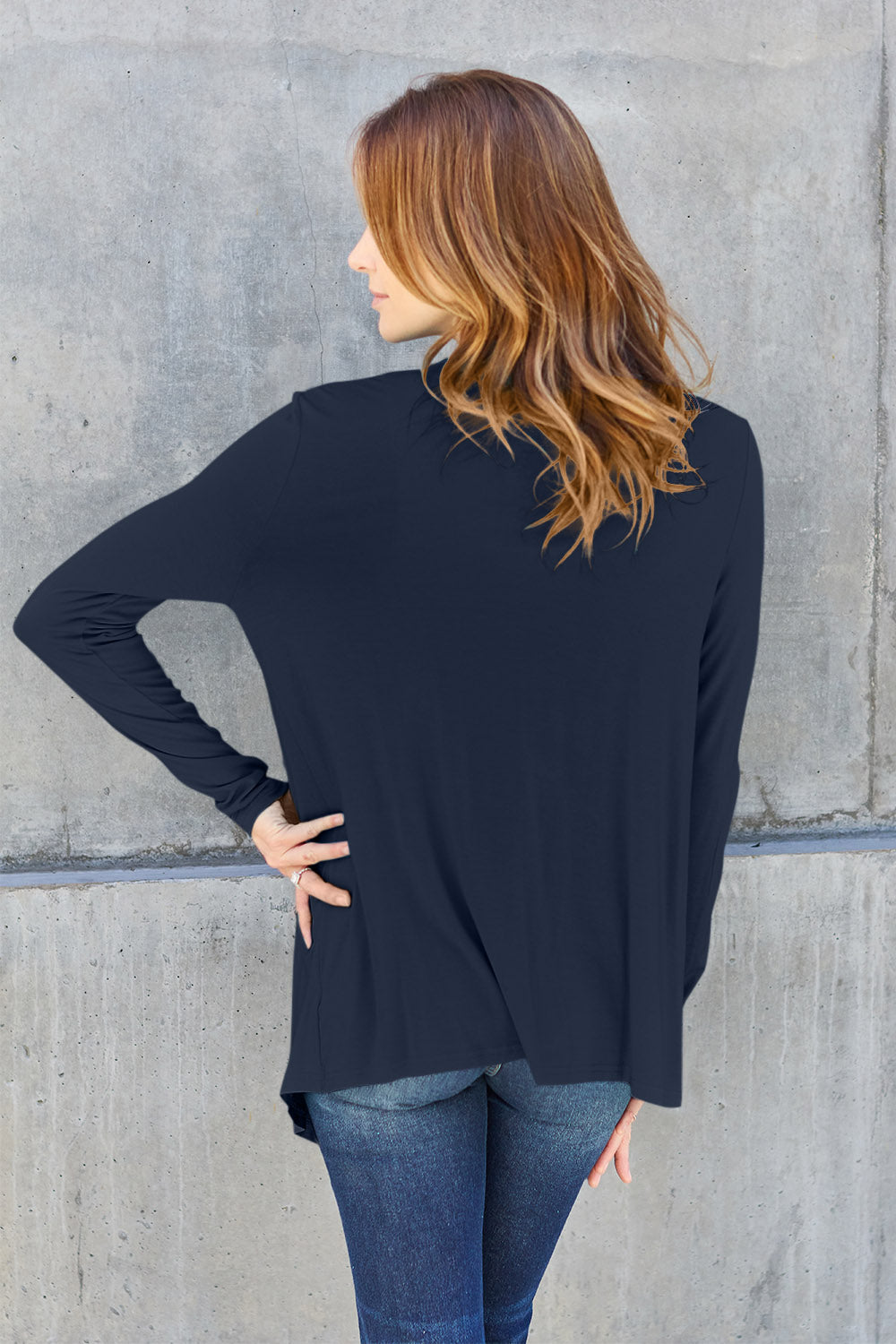 A woman wearing the Basic Bae Full Size Open Front Long Sleeve Cardigan in navy blue, paired with a white top and blue jeans, stands against a concrete wall. She touches her hair and gazes to the side, embodying a chic yet simple style. The slightly stretchy materials of the cardigan ensure comfort while maintaining its stylish simplicity. For easy care, machine wash it cold.