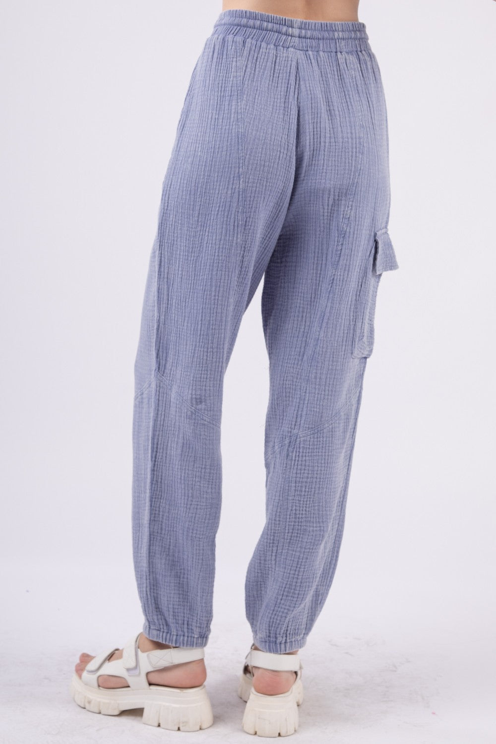 A person wearing VERY J Washed Woven Crinkle Gauze Drawstring Cargo Pants in light blue, paired with white sandals. These relaxed-fit, textured pants feature an elastic waistband and pockets, showcasing a utility design that's perfect for blending comfort and functionality against a plain background.