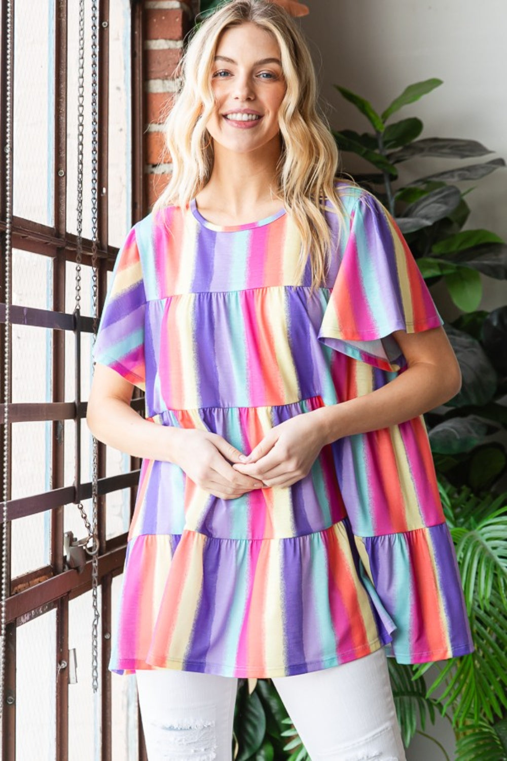 A person wearing the Heimish Full Size Short Sleeve Striped Tiered Top, featuring a colorful, classic striped pattern and flared sleeves, stands by a window. They are smiling, surrounded by plants in the background.