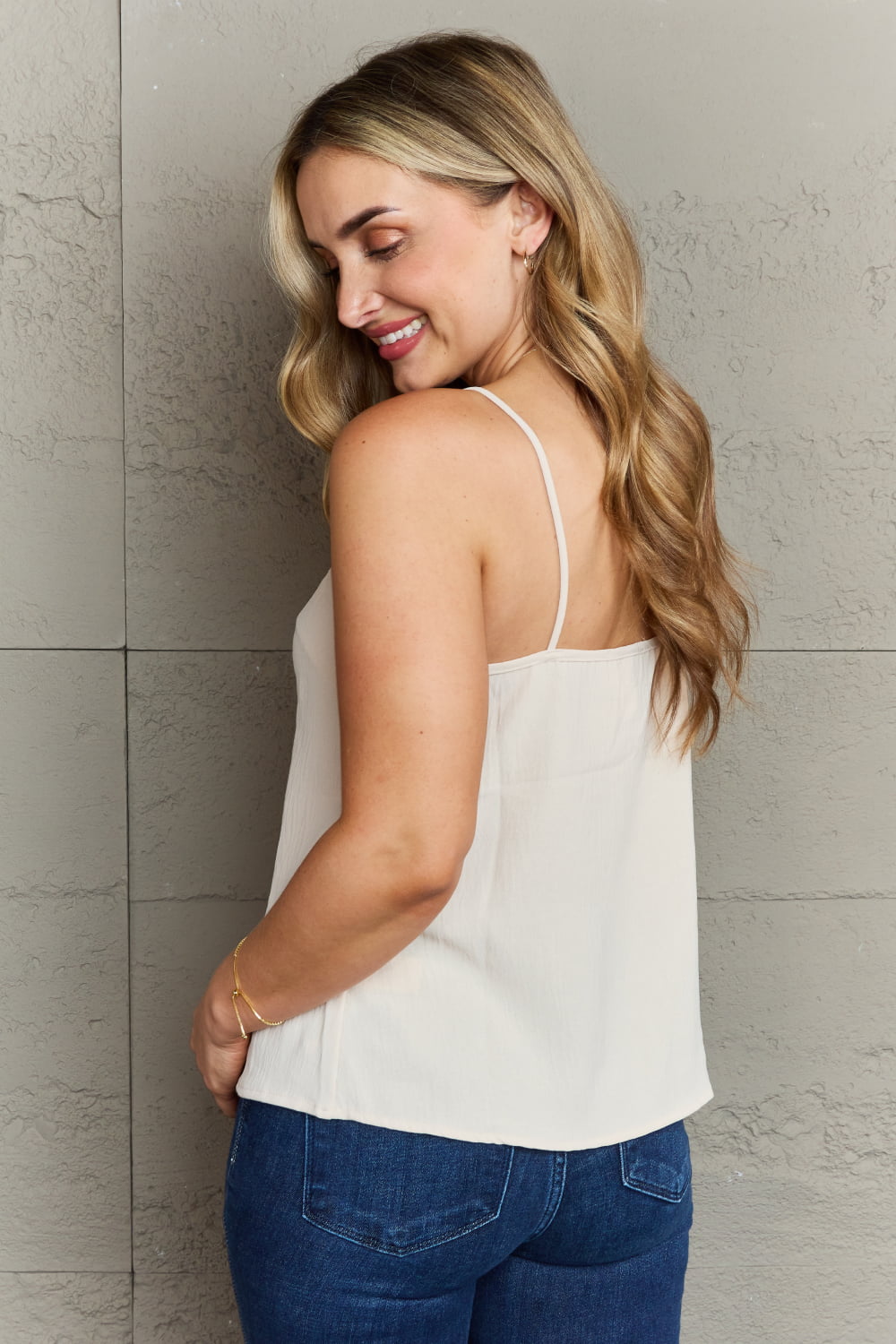 A person with long wavy hair wearing a Ninexis For The Weekend Loose Fit Cami and blue jeans stands against a gray textured wall.