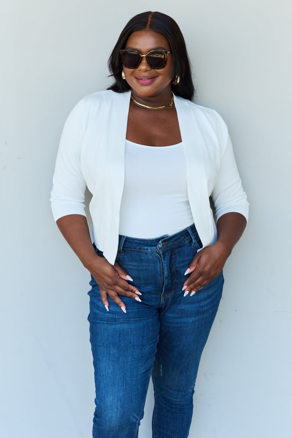 A stylish person wearing sunglasses, a Doublju My Favorite Full Size 3/4 Sleeve Cropped Cardigan in Ivory, and blue jeans poses in front of a plain background, effortlessly highlighting their versatile look.