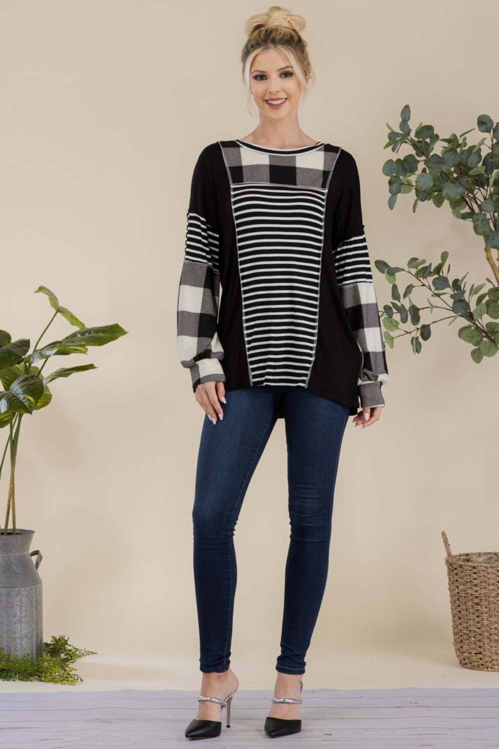 A person wearing a Celeste Full Size Color Block Exposed Seam T-Shirt, featuring black and white plaid with reverse stitching and long sleeves, poses while smiling in front of a neutral background with a plant.