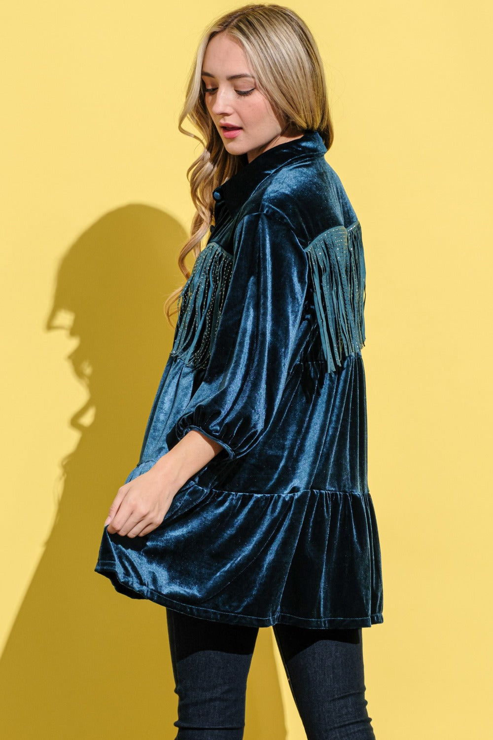 A woman in the And The Why Fringe Detailed Velvet Shirt Dress with a tailored fit and button-up front, wearing jeans, stands against a yellow background.