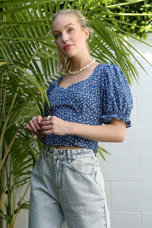 A person wearing the SS puff sleeve top, featuring a blue floral print, pairs it with light jeans while standing on grass beside potted plants, against a white wall backdrop.