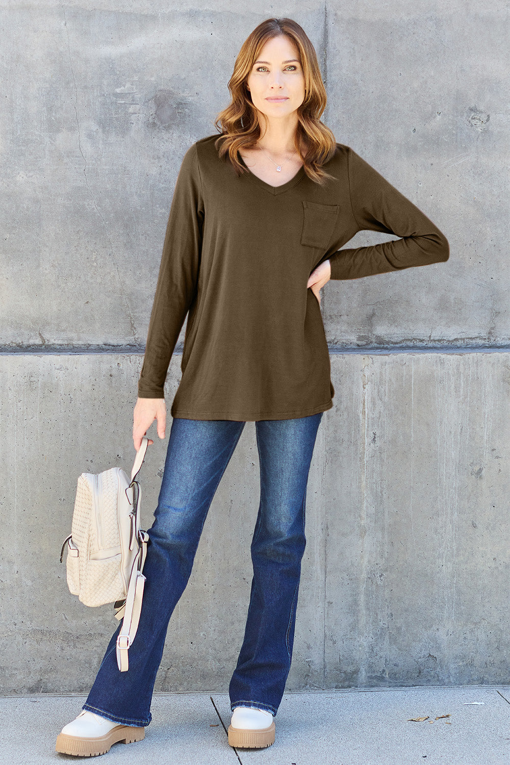 A woman with light brown hair stands against a concrete wall, wearing the Basic Bae Full Size V-Neck Long Sleeve Top in olive green and blue jeans, with a white backpack on her shoulder. Her outfit exudes basic style and is made from polyester, ensuring it’s both comfortable and suitable for machine wash cold. She is looking at the camera.