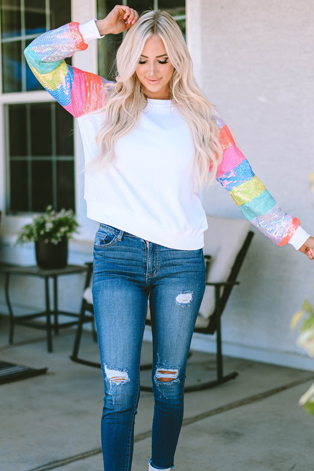 A person with long blonde hair is seen from the back, wearing a White Sequin Color Block Raglan Sleeve Pullover Sweatshirt and blue jeans. They are standing in front of a building with large windows.