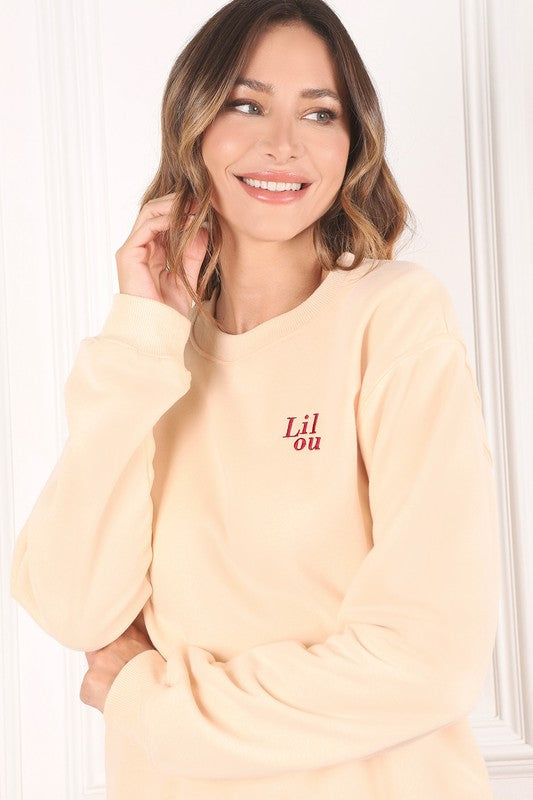 A person wearing a cream sweatshirt with embroidered "Lilou" logo is standing against a white background.