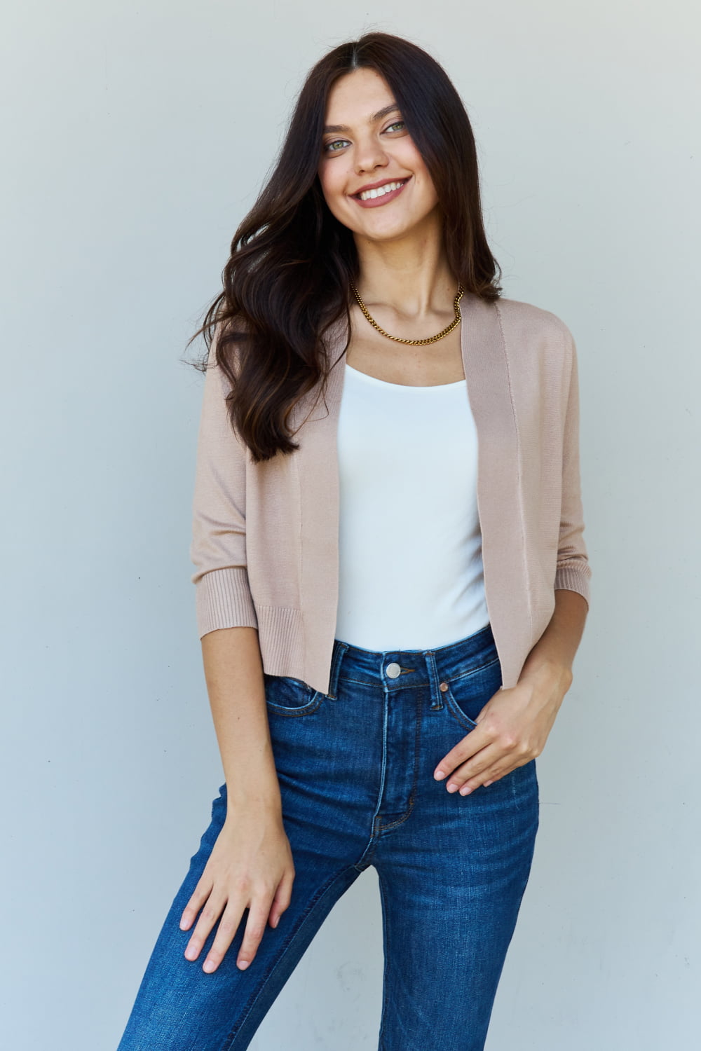 Woman with long dark hair wearing the versatile Doublju My Favorite Full Size 3/4 Sleeve Cropped Cardigan in Khaki over a white top and blue jeans, standing against a plain background.