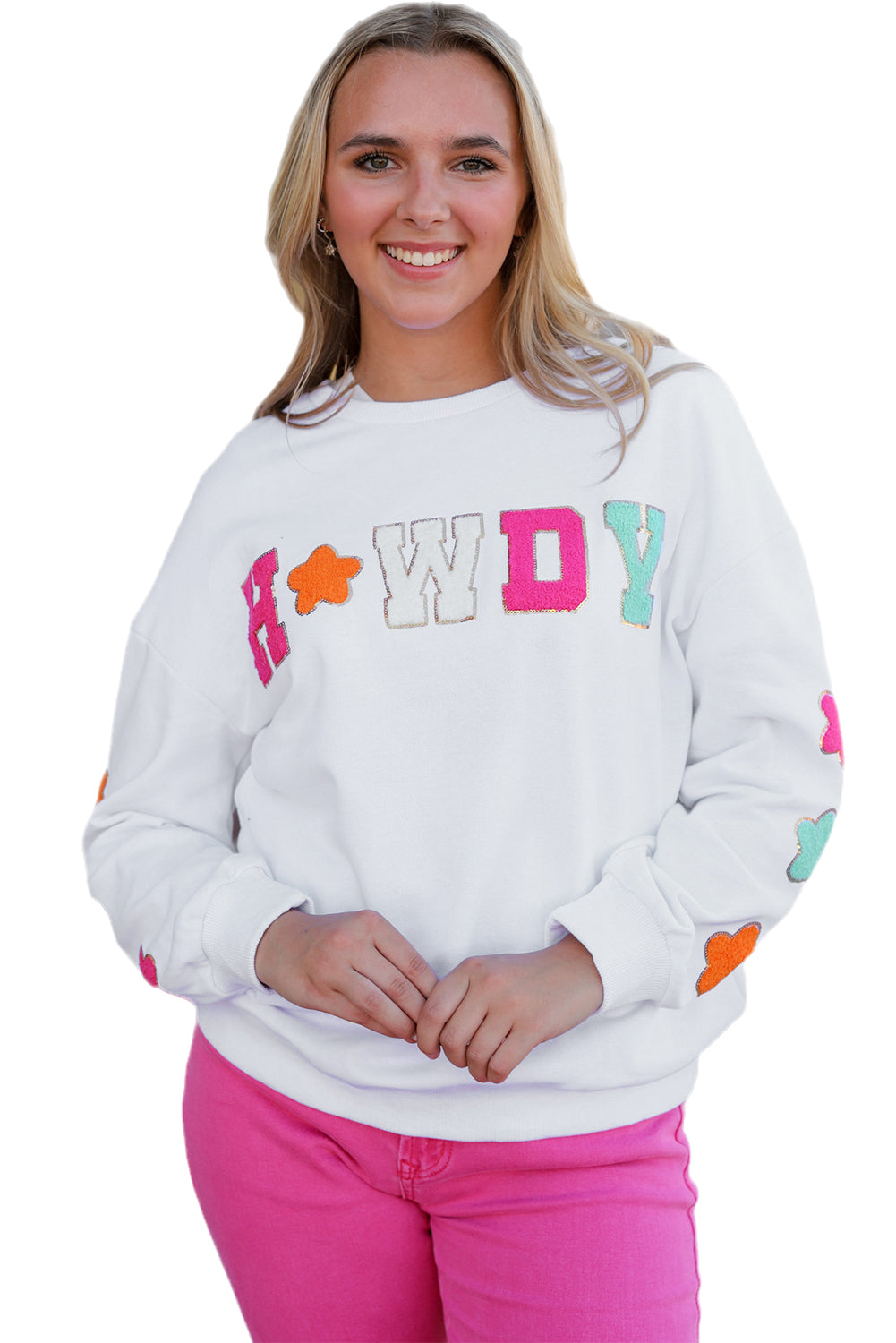 A person poses in the White Howdy Glitter Chenille Patch Graphic Casual Sweatshirt, adorned with colorful star patterns and the letters "WD." She wears light denim shorts and has shoulder-length blonde hair.