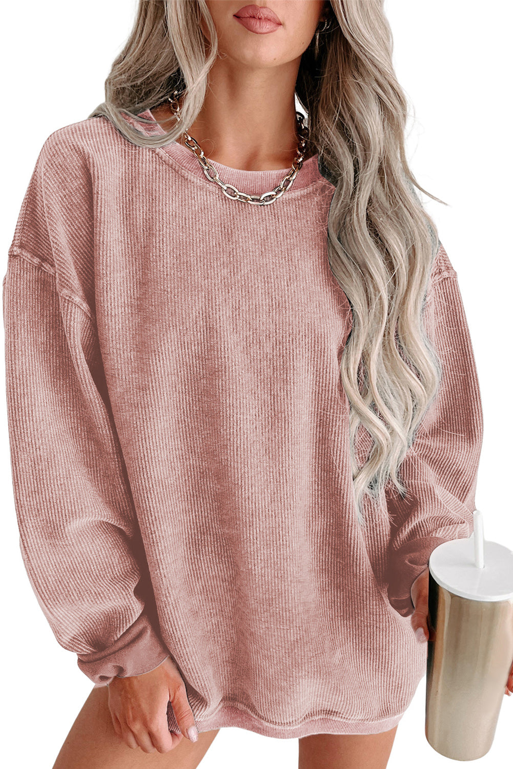 A woman with long, wavy hair is sitting on a high stool, smiling and holding a tumbler. She is wearing a relaxed fit Pink Solid Ribbed Knit Round Neck Pullover Sweatshirt, white shorts, a chain necklace, and knee-high white boots.