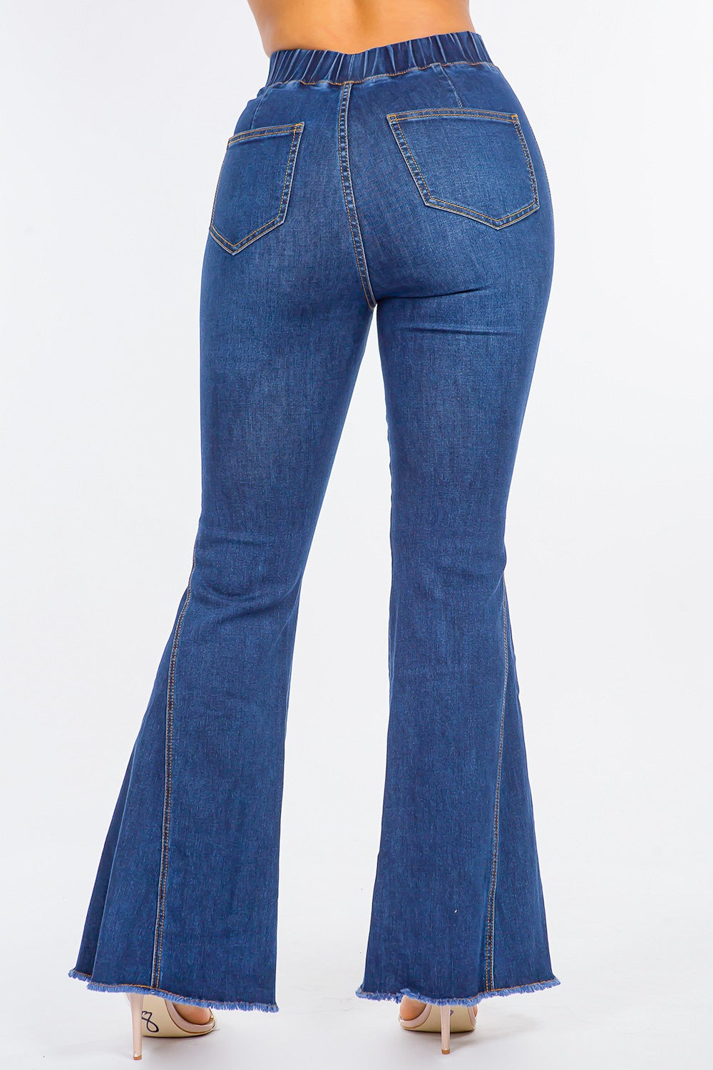 The image shows a person wearing American Bazi High Waist Curvy Flare Jeans made from stretchy denim, paired with nude open-toe heels. The jeans feature an elastic waistband and frayed hems, adding a touch of retro flair. The background is plain white.