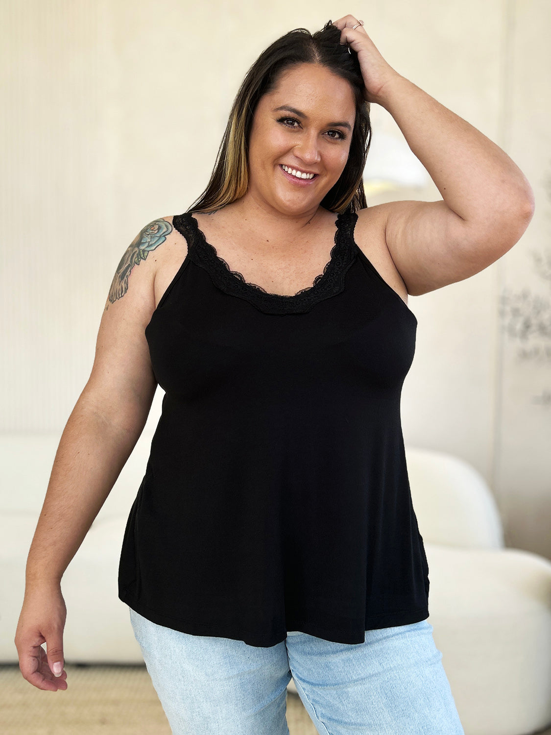 A woman with long hair wearing sunglasses, a slightly stretchy pink "Basic Bae Full Size Lace Detail V-Neck Cutout Cami," and basic style blue jeans stands against a concrete wall, looking to the side with one hand touching her upper chest.