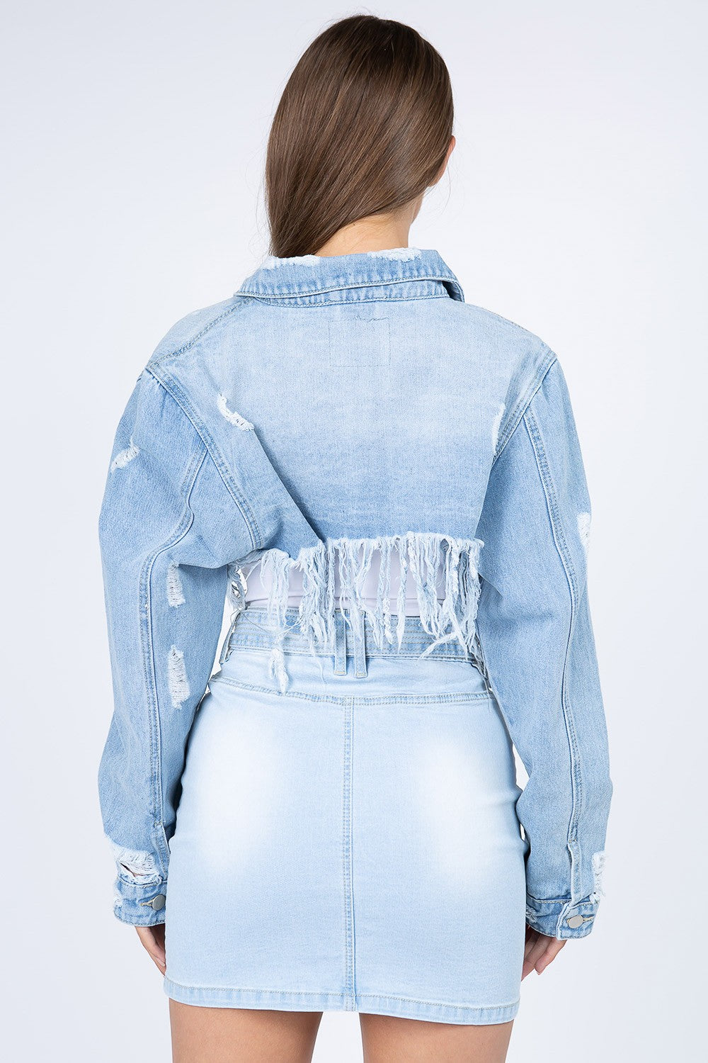 A woman with long brown hair wears an American Bazi Distressed Denim Jacket with Frayed Hem and a light denim skirt with front pockets. The jacket features frayed hems, adding a touch of edgy style. She stands facing the camera against a plain white background.