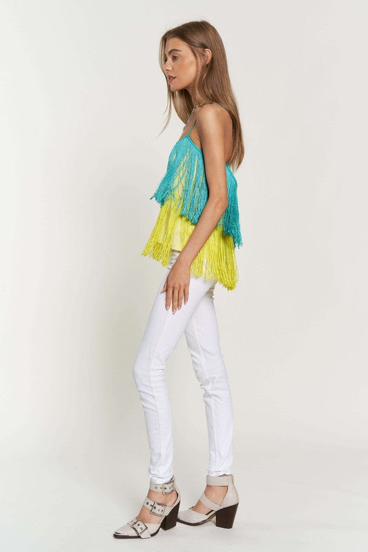 A woman with long brown hair is wearing a Fringe Overlay Cross Straps Party Cami Top featuring blue and yellow layers, paired with white pants, standing against a plain background.