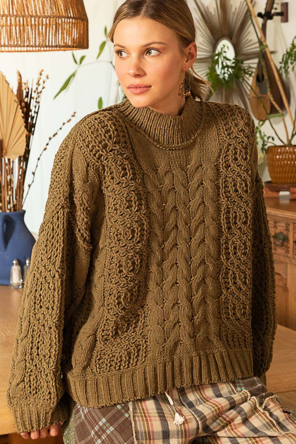 A person sits on a patterned blanket, wearing the POL Mock Neck Cable Knit Sweater and patterned pants, with plants and decorative items in the background.