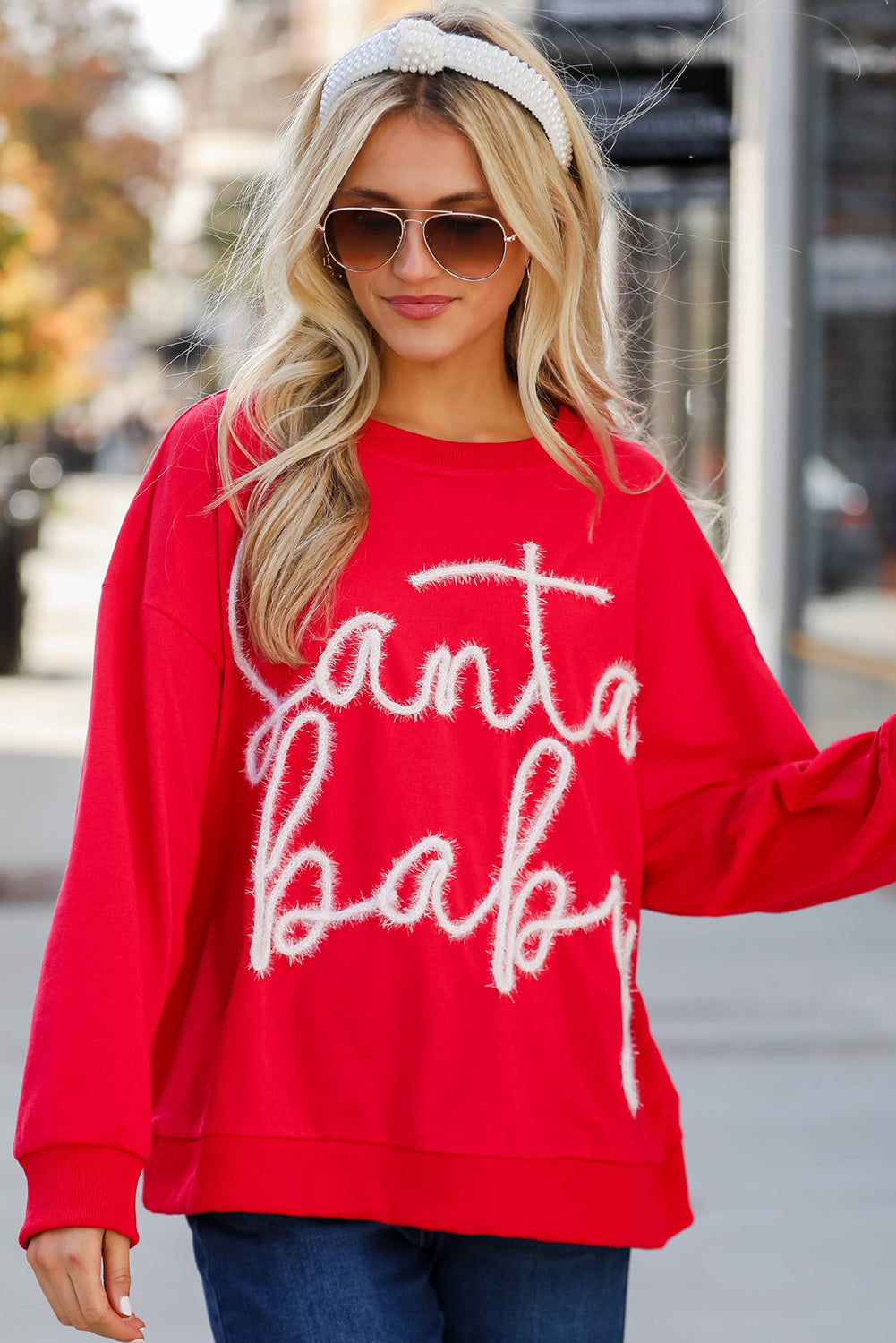 A person with long blonde hair is seen wearing the Fiery Red Christmas Santa Baby Tinsel Graphic Sweatshirt and blue jeans, facing away from the camera against a plain background.