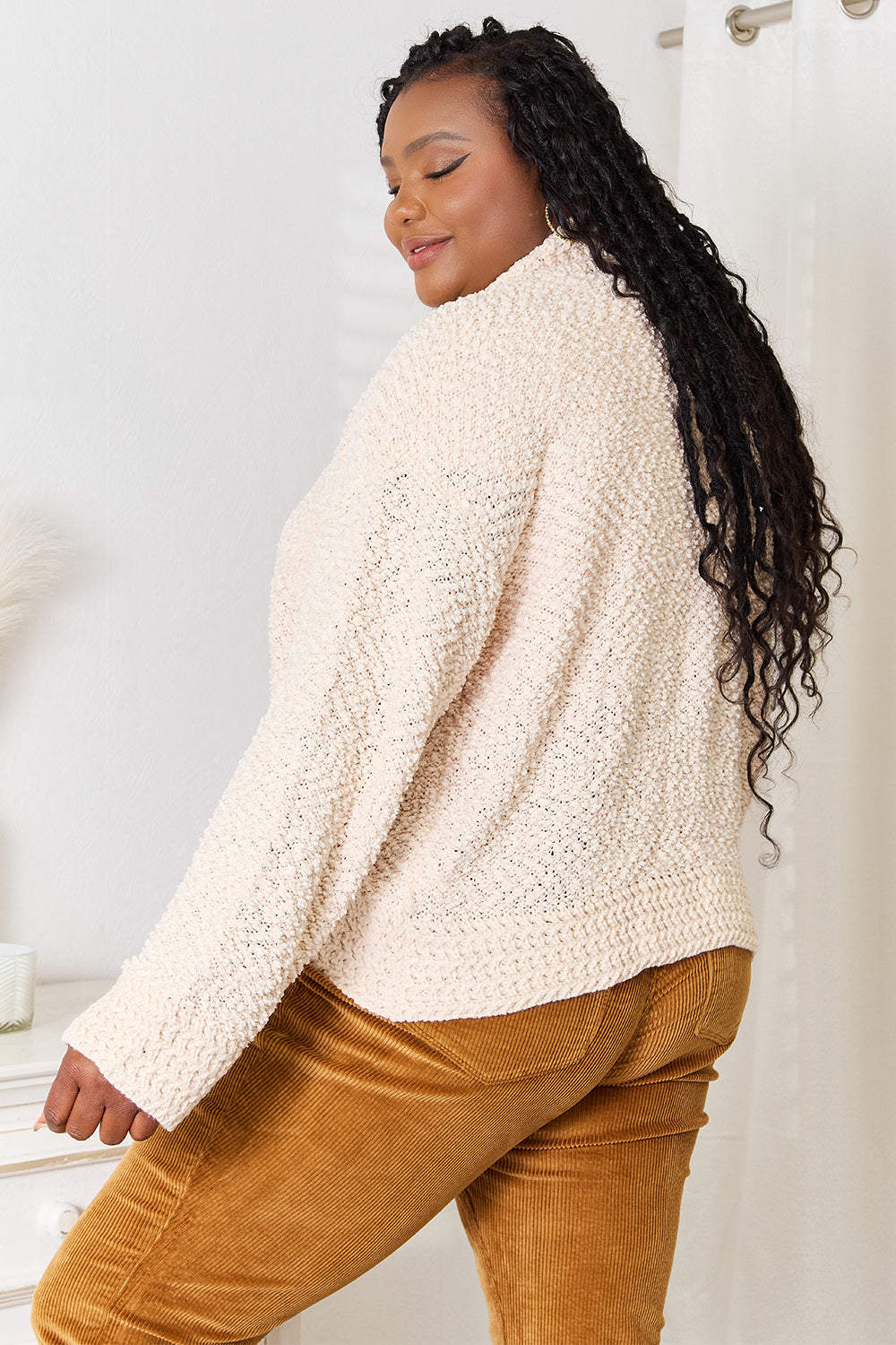 A person with long braided hair is standing and wearing the Double Take Open Front Long Sleeve Cardigan in beige over a white top, paired with mustard-colored pants. They are looking to the side, holding the comfortable fabric of the cardigan together.