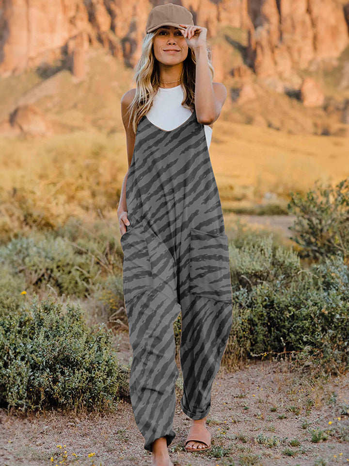 A person wearing a casual white V-neck tank top and a Full Size Printed V-Neck Sleeveless Jumpsuit with a brown animal print zebra-stripe pattern stands outdoors with one hand on their hat and the other in their pocket, set against a rocky landscape backdrop.