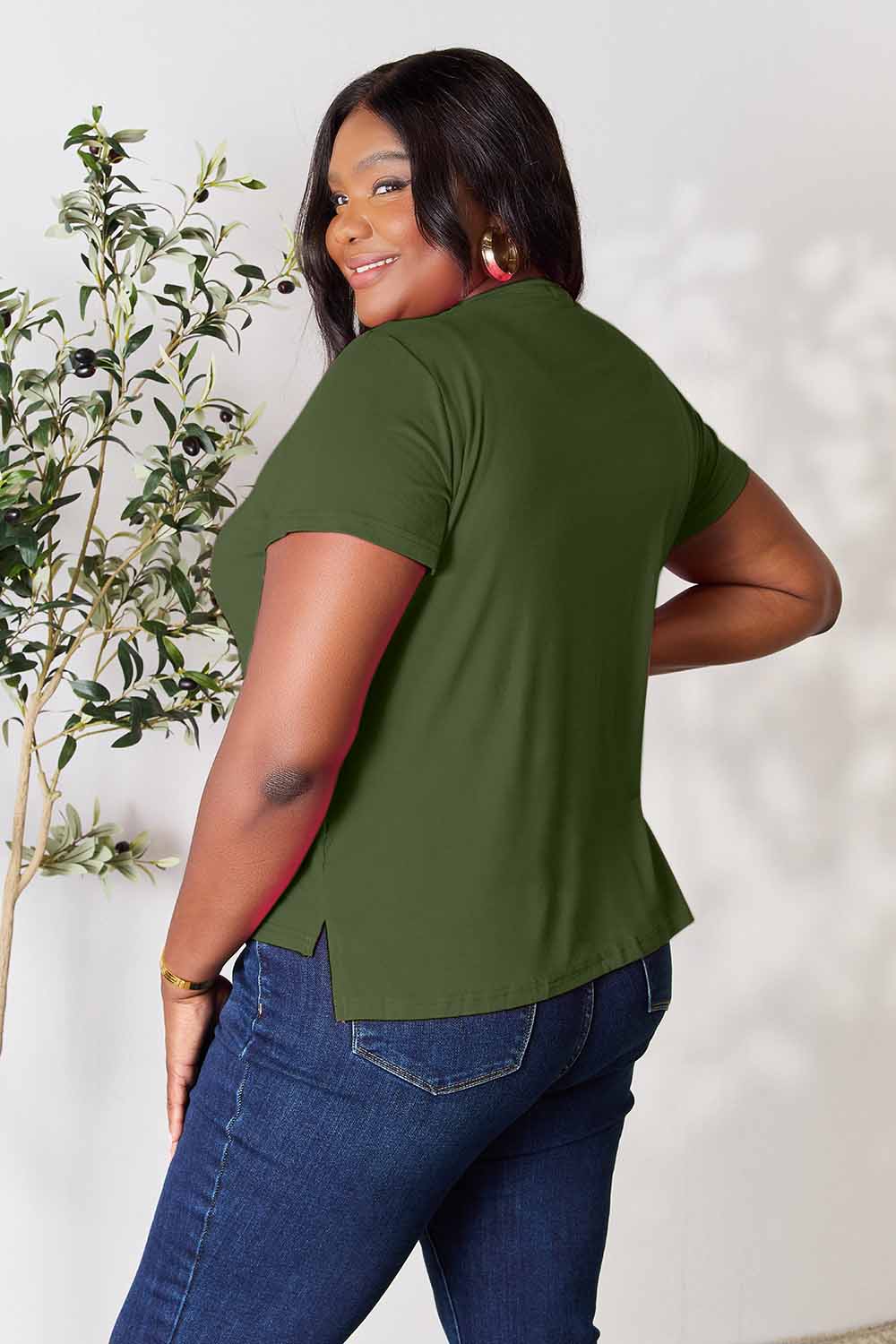 A woman stands next to an indoor plant, smiling, with one hand in her khaki pants pocket. In her pink Basic Bae Full Size Round Neck Short Sleeve T-Shirt and gold jewelry, she radiates casual elegance.