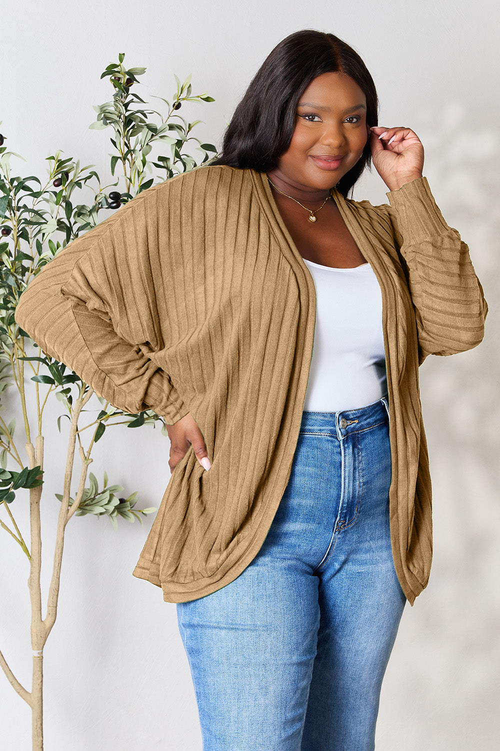 A person with wavy hair, wearing the Basic Bae Full Size Ribbed Cocoon Cardigan over a white top and beige pants, stands in front of a light-colored background with a leafy plant, showcasing an effortlessly basic style.