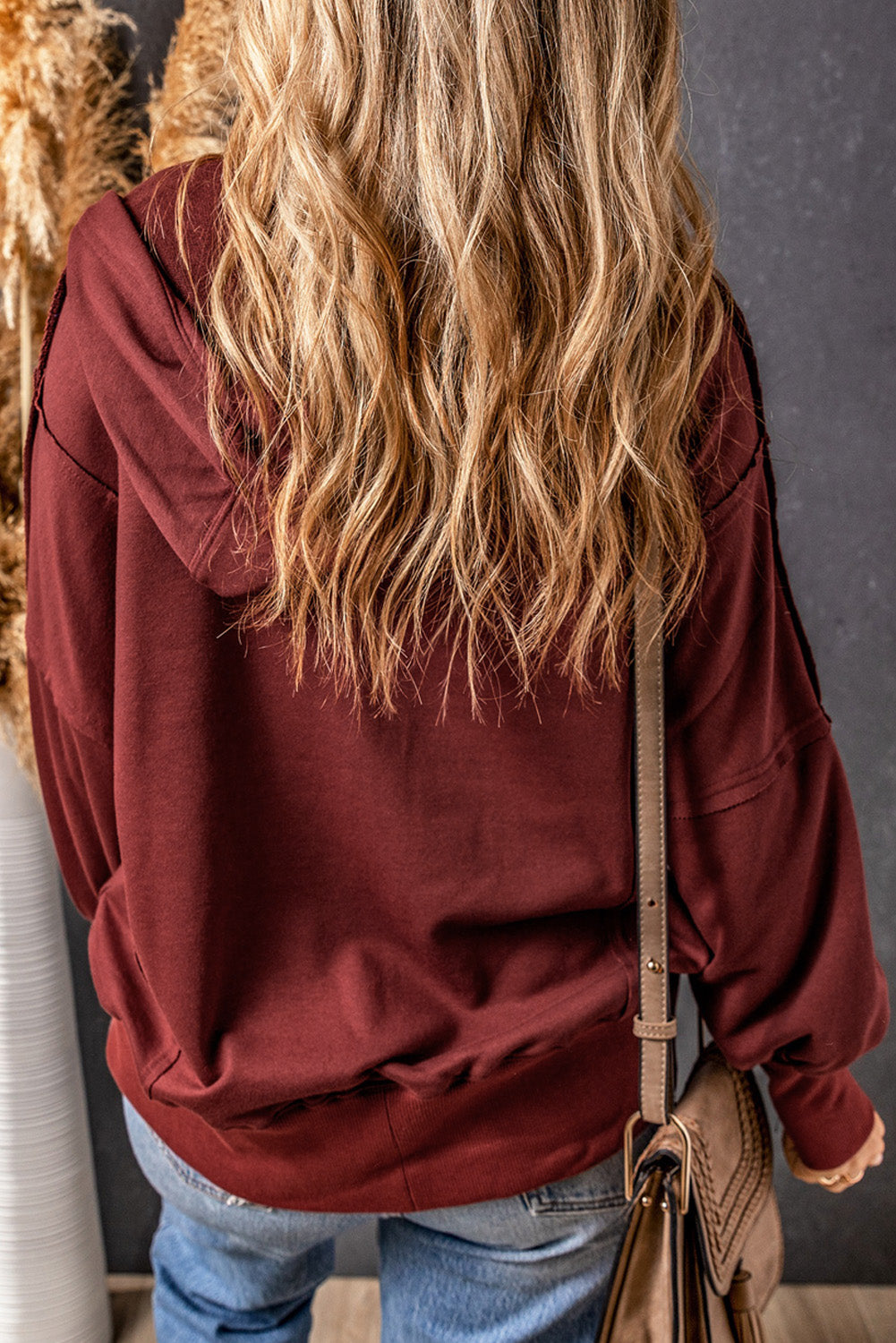 A woman with long, wavy blonde hair is seen from behind, wearing a trendy Brown Raw Edge Exposed Seam Full Zip Hoodie and carrying a brown shoulder bag.