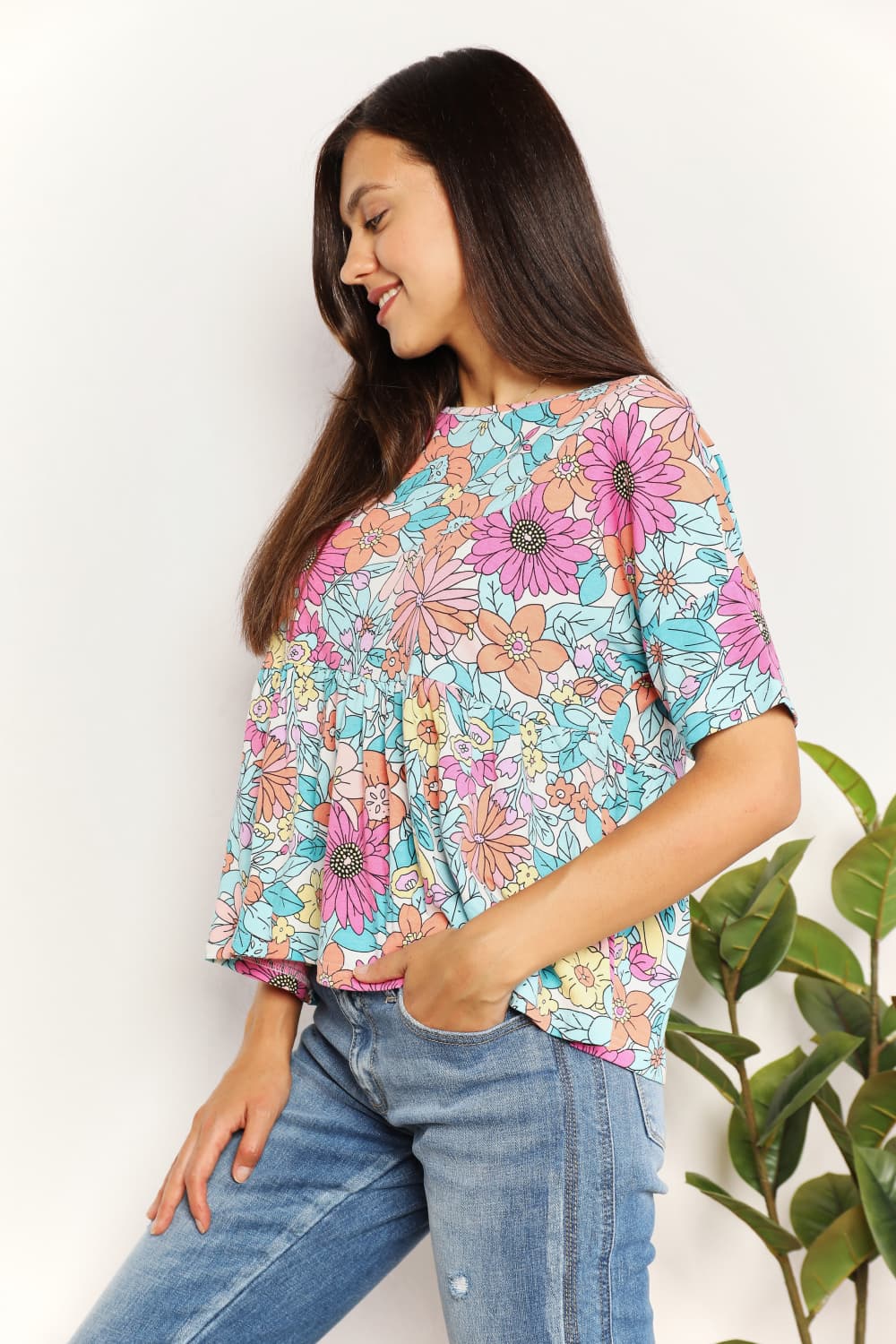 A person wearing a Double Take Floral Round Neck Babydoll Top and jeans stands with one hand in their pocket, with a plant in the background.