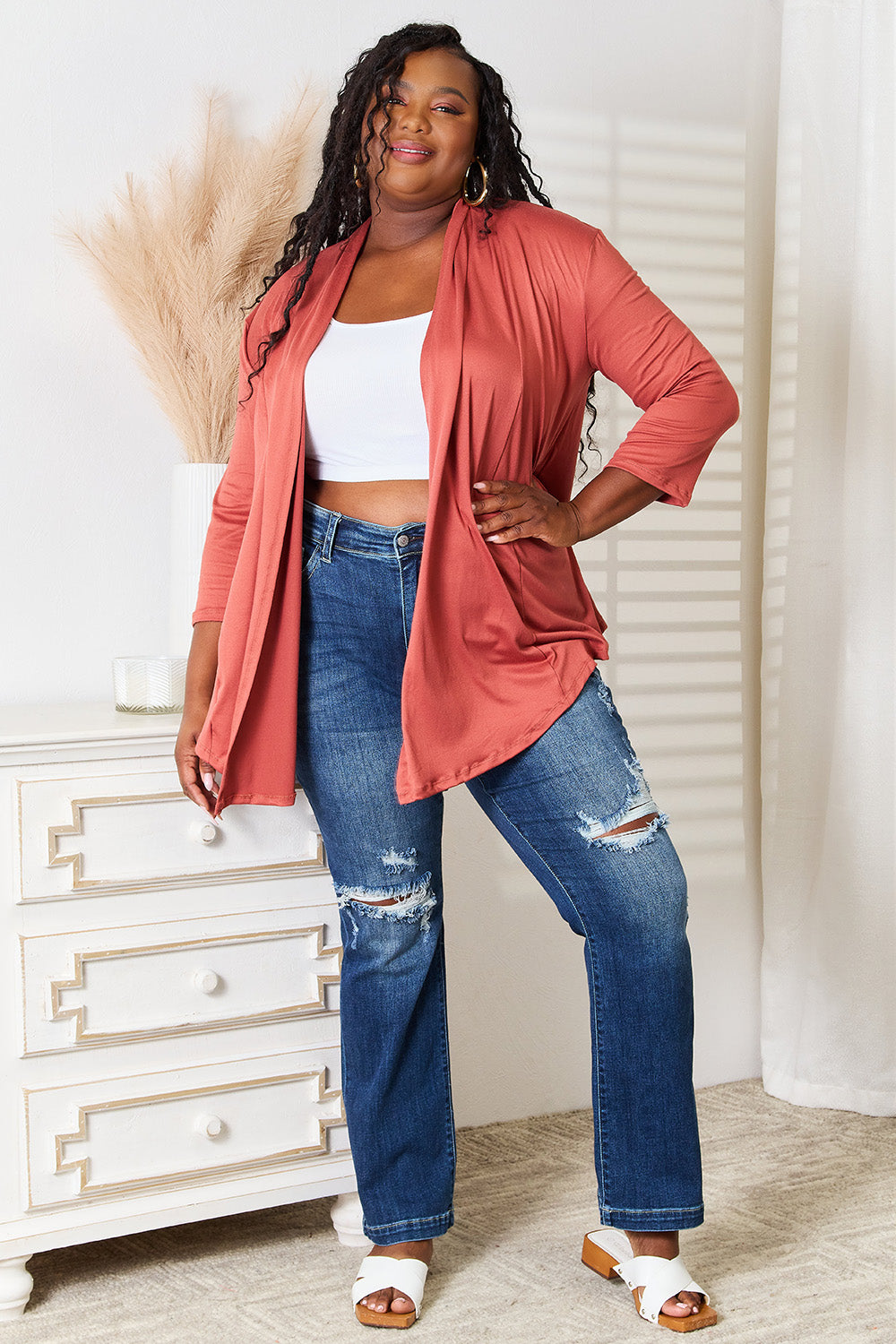 A person with long hair is wearing the Culture Code Full Size Open Front Cardigan in a rust color, complemented by a flattering draped front. They pair it with a white top and ripped jeans while standing indoors with one hand on their hip.