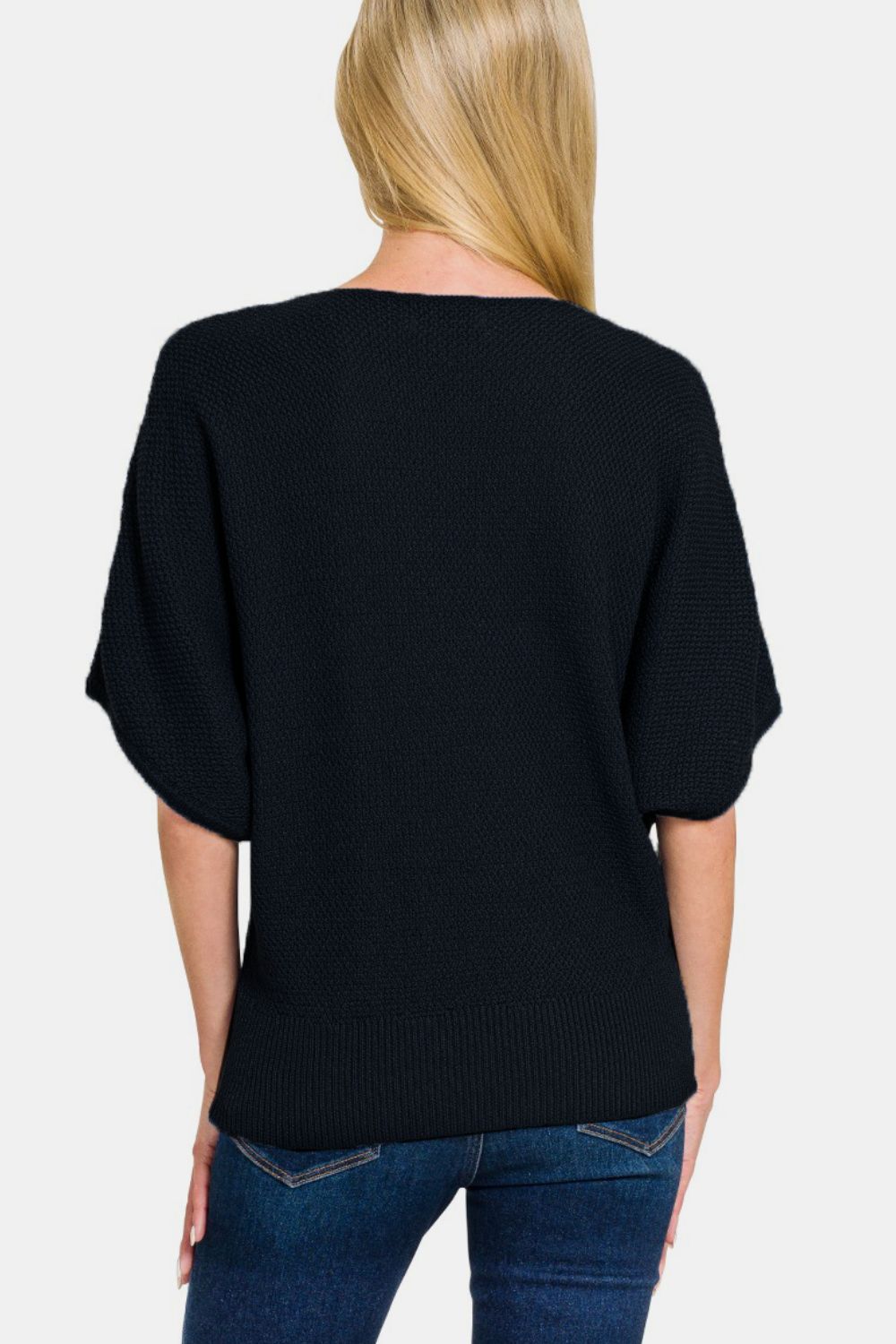 A person with long, blonde hair is wearing a Zenana V-Neck Short Sleeve Dolman Sweater in black and blue jeans, standing against a plain white background.