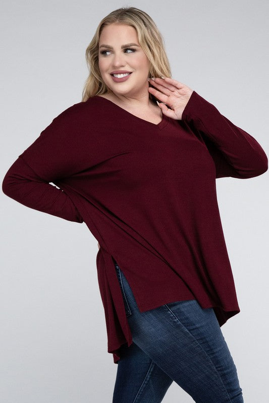 A woman with long, blonde hair wears a Plus Dolman Sleeve V-Neck Side Slit Hi-Low Hem Top and blue jeans, posing with one hand on her hip and smiling at the camera.