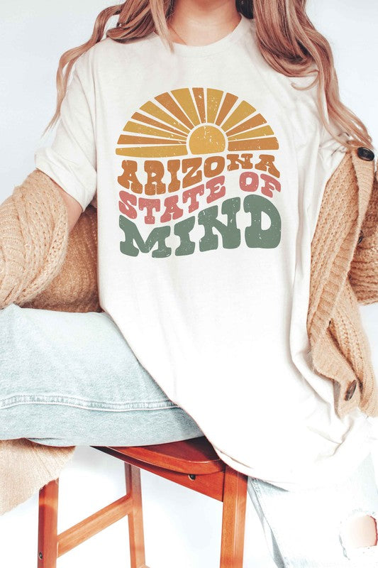 Person wearing the ARIZONA STATE OF MIND Graphic Tee, a beige 100% cotton t-shirt with "ARIZONA STATE OF MIND" text in brown and green colors. The unisex shirt offers both comfort and style for any occasion. They are holding a pink polka-dotted mug.