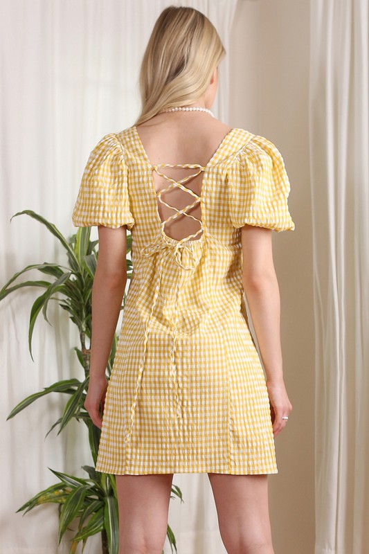 A woman stands indoors, smiling while wearing the SS back strap dress in yellow gingham, and holding a white purse.