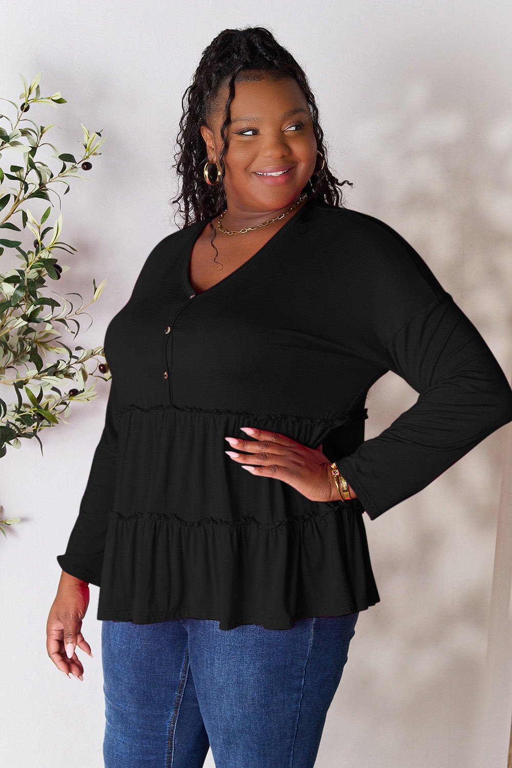 Person stands indoors near a plant, wearing the Double Take Half Button Long Sleeve Ruffle Hem Blouse in gray and blue jeans, smiling while looking to the side. The slightly stretchy fabric of the blouse adds comfort while its buttoned design offers a touch of elegance.