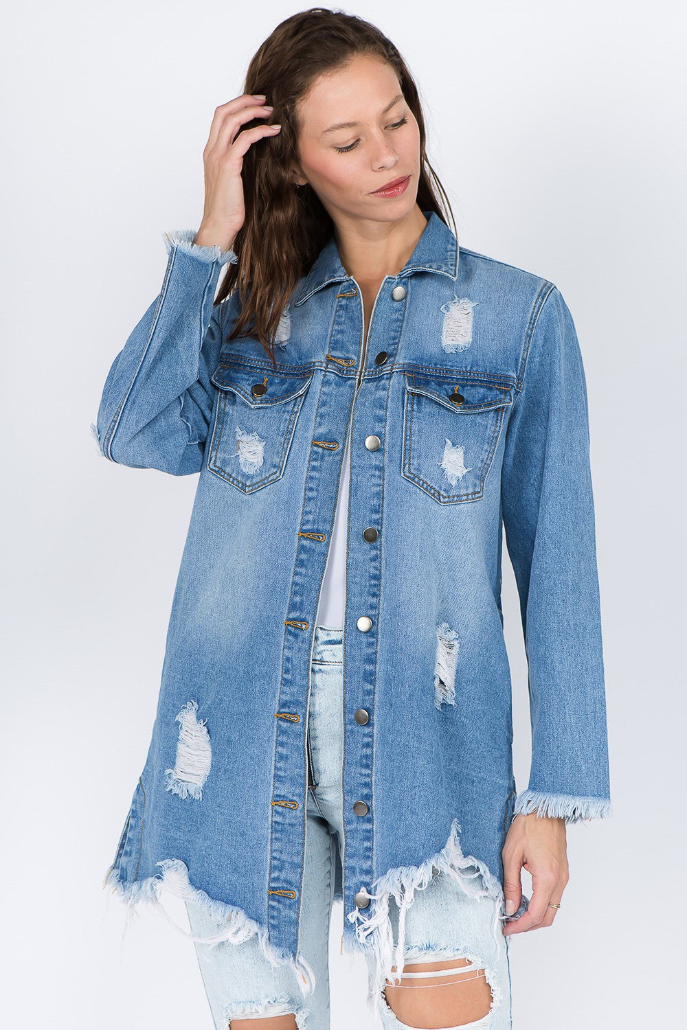 A woman models the American Bazi Distressed Frayed Hem Denim Jacket, featuring frayed edges, a button-up front, and chest pockets that add to its vintage feel. She has long hair and is looking downward with one hand touching her head.