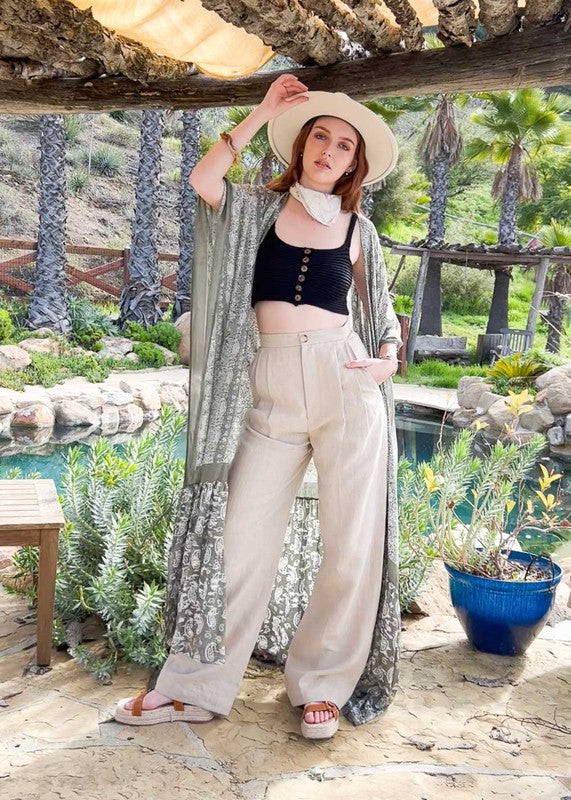 A woman wearing a Paisley Tapestry Free Flow Kimono stands in a bright, eclectic room with plants, books, and an oversized deer head on the wall. The space features large windows and industrial decor.