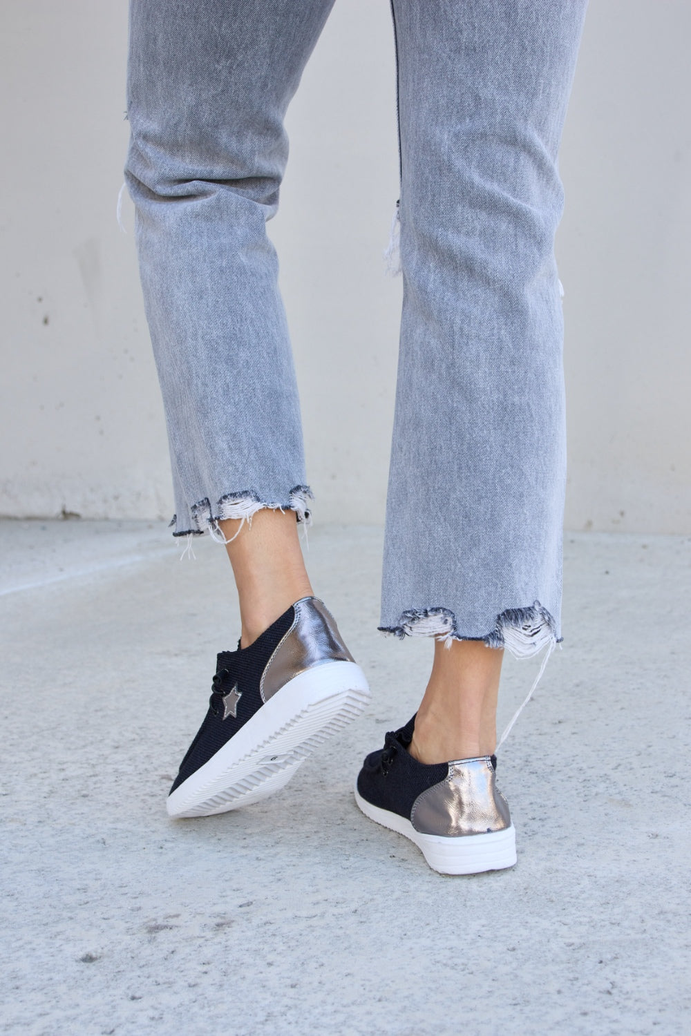 A person stands on a concrete surface, wearing distressed grey jeans and stylish Forever Link Star Lace-Up Flat Sneakers featuring black uppers and white soles.