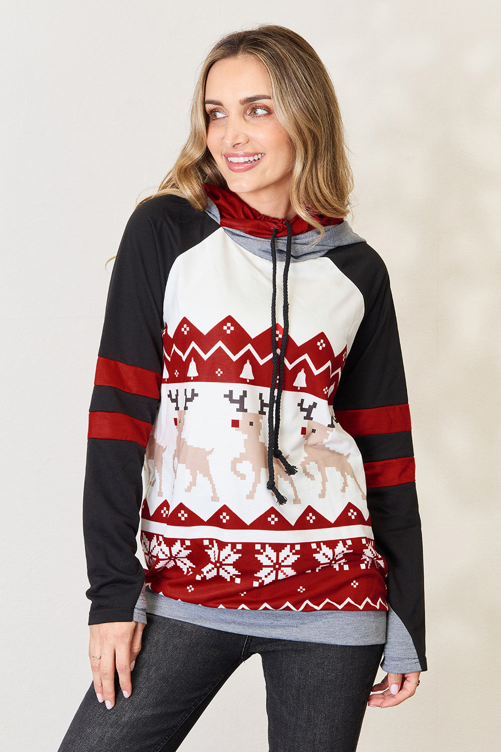 A person is wearing the Double Take Full Size Christmas Drawstring Long Sleeve Hoodie, which features a holiday-themed red and white pattern with reindeer and snowflakes. This cozy hoodie, made from a polyester spandex blend, has stylish black sleeves accented with red stripes. The person is smiling and looking to the side.