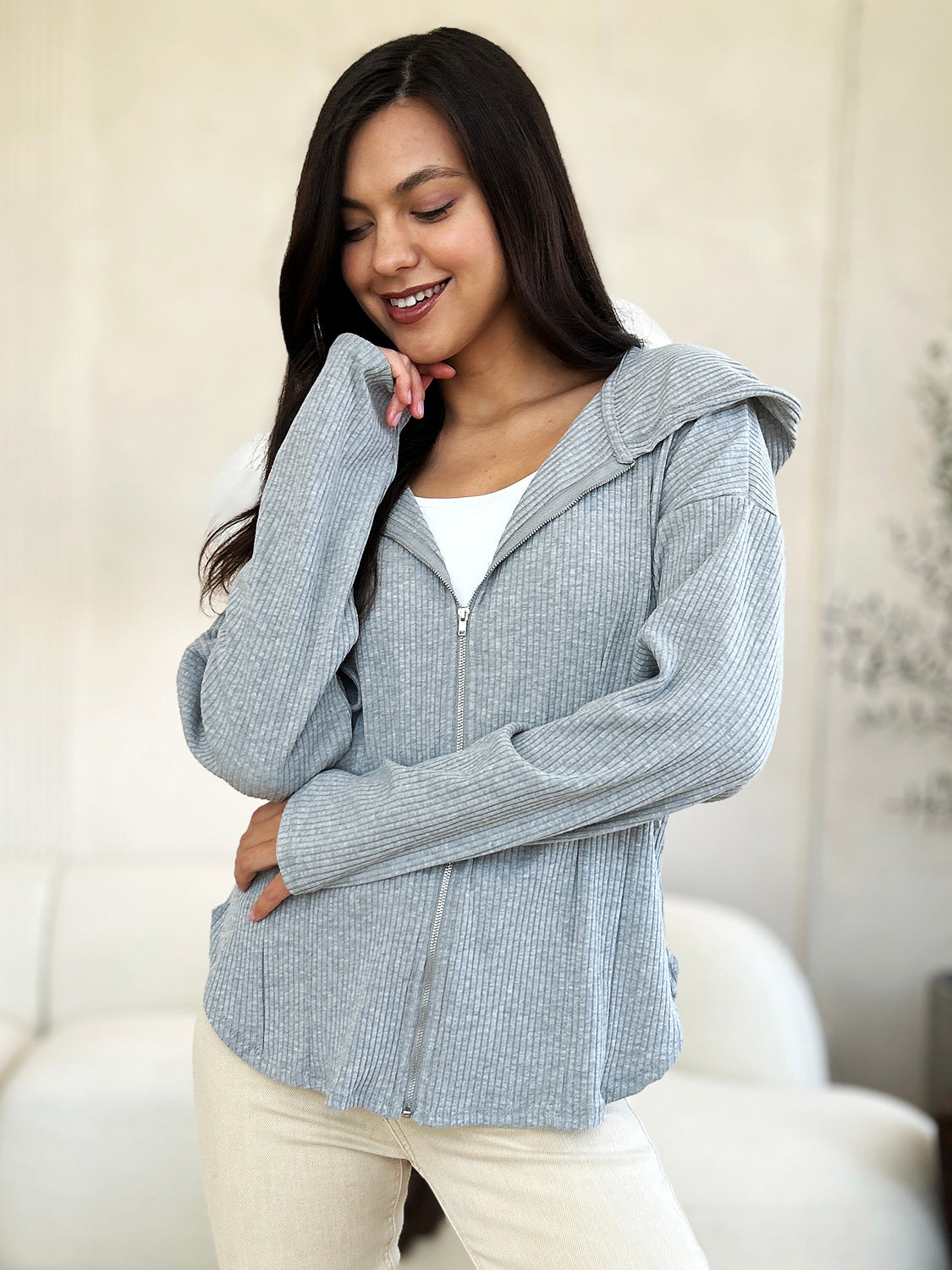 A woman in a trendy Culture Code Full Size Ribbed Zip Up Drawstring Hooded Jacket and light-colored pants stands indoors, smiling with her eyes closed and one hand resting on her chin.